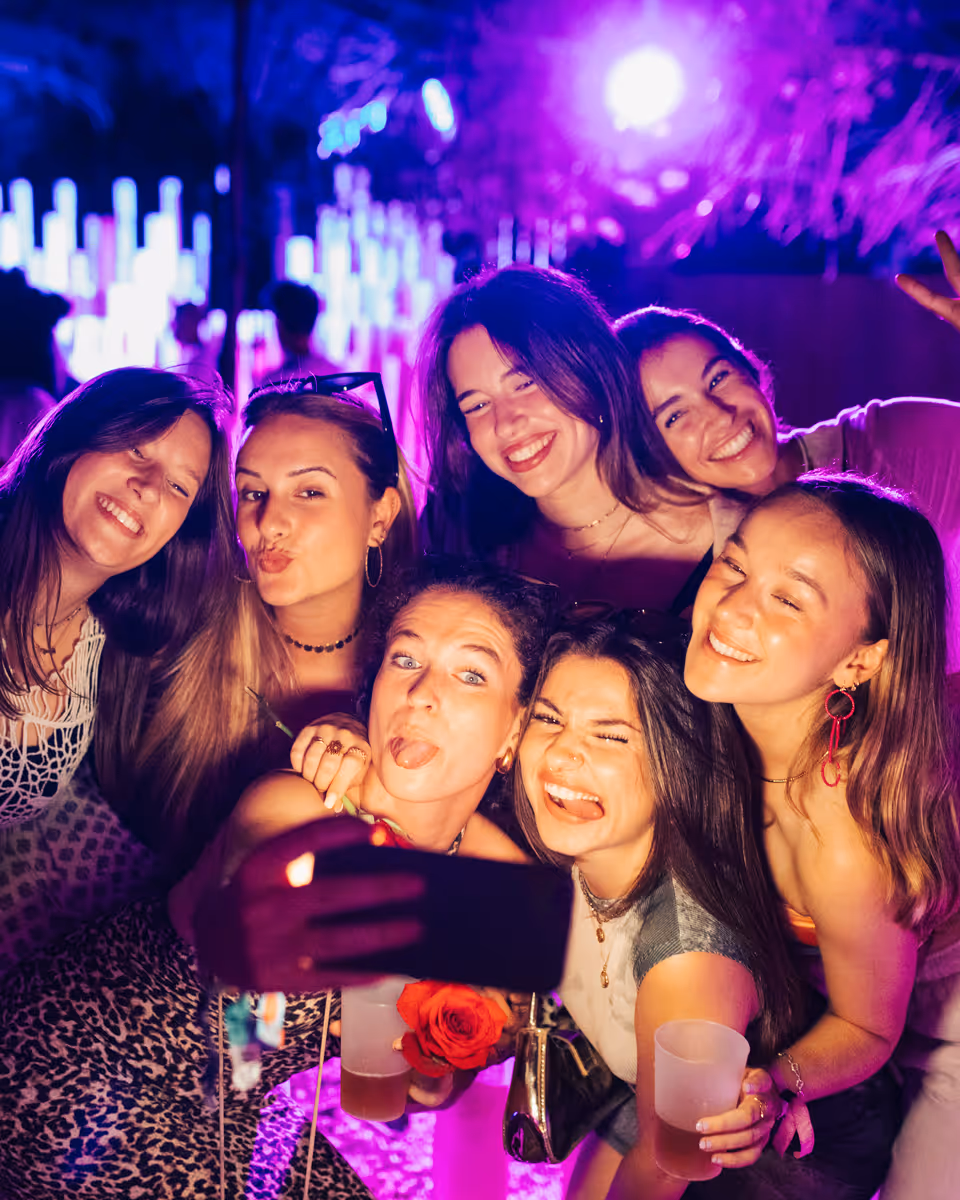 Group of seven young women smiling and posing closely for a selfie at a vibrant party with purple lighting.