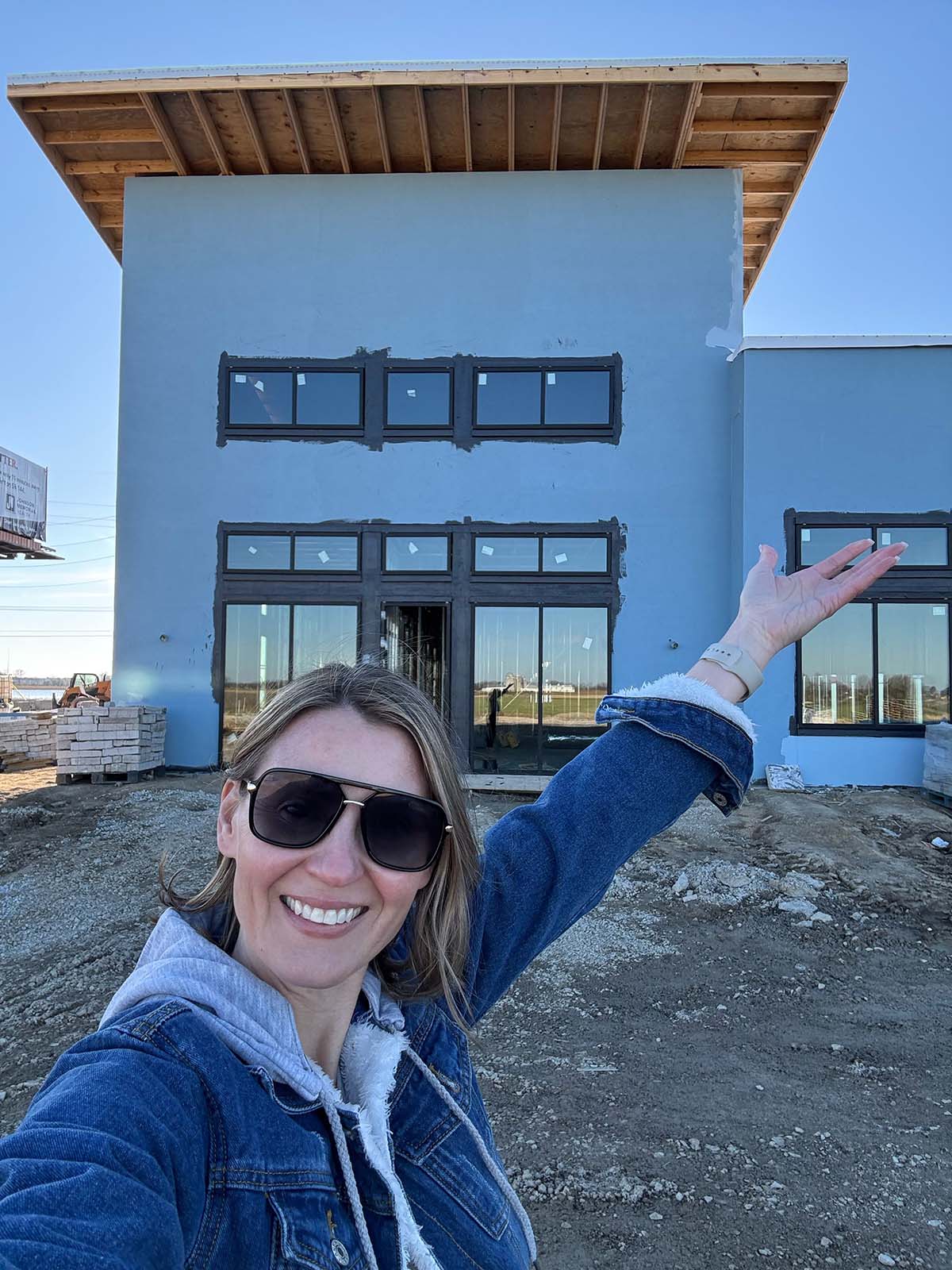 Smiling woman in sunglasses and denim jacket gestures to a newly built modern house exterior on a clear day.
