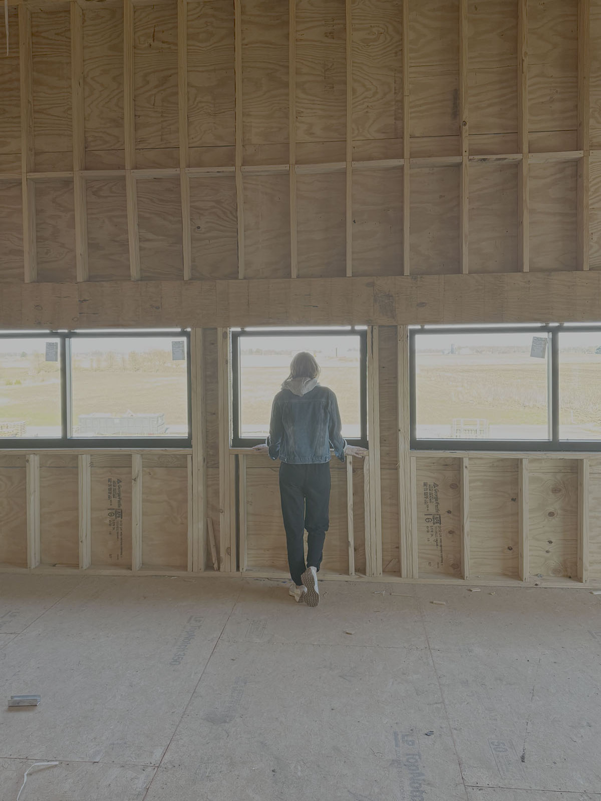 Person in denim jacket standing inside unfinished wooden building looking out three framed windows.
