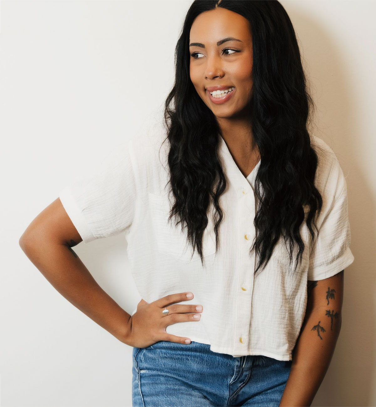 Smiling woman with long black hair, wearing a white button-up shirt and blue jeans, hand on hip.