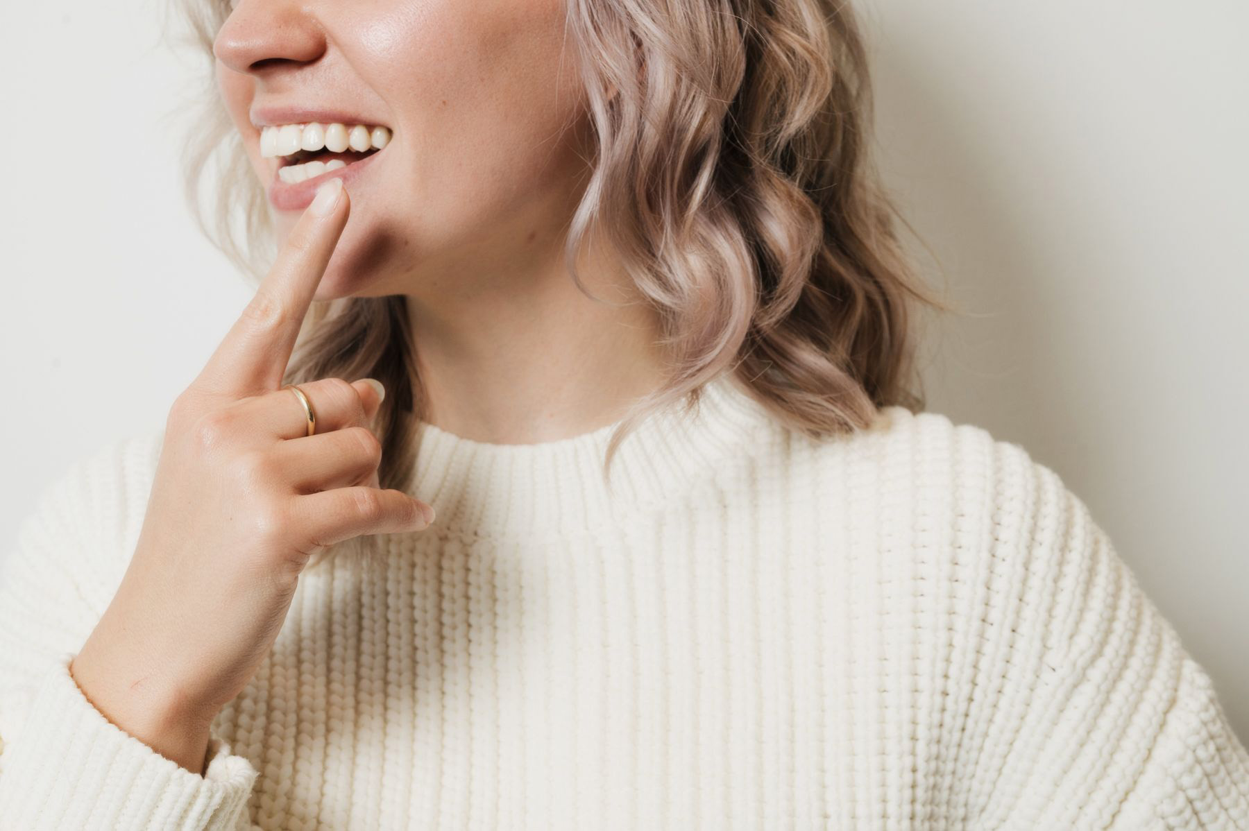 Smiling woman with wavy hair touching her face with her finger wearing a white sweater.