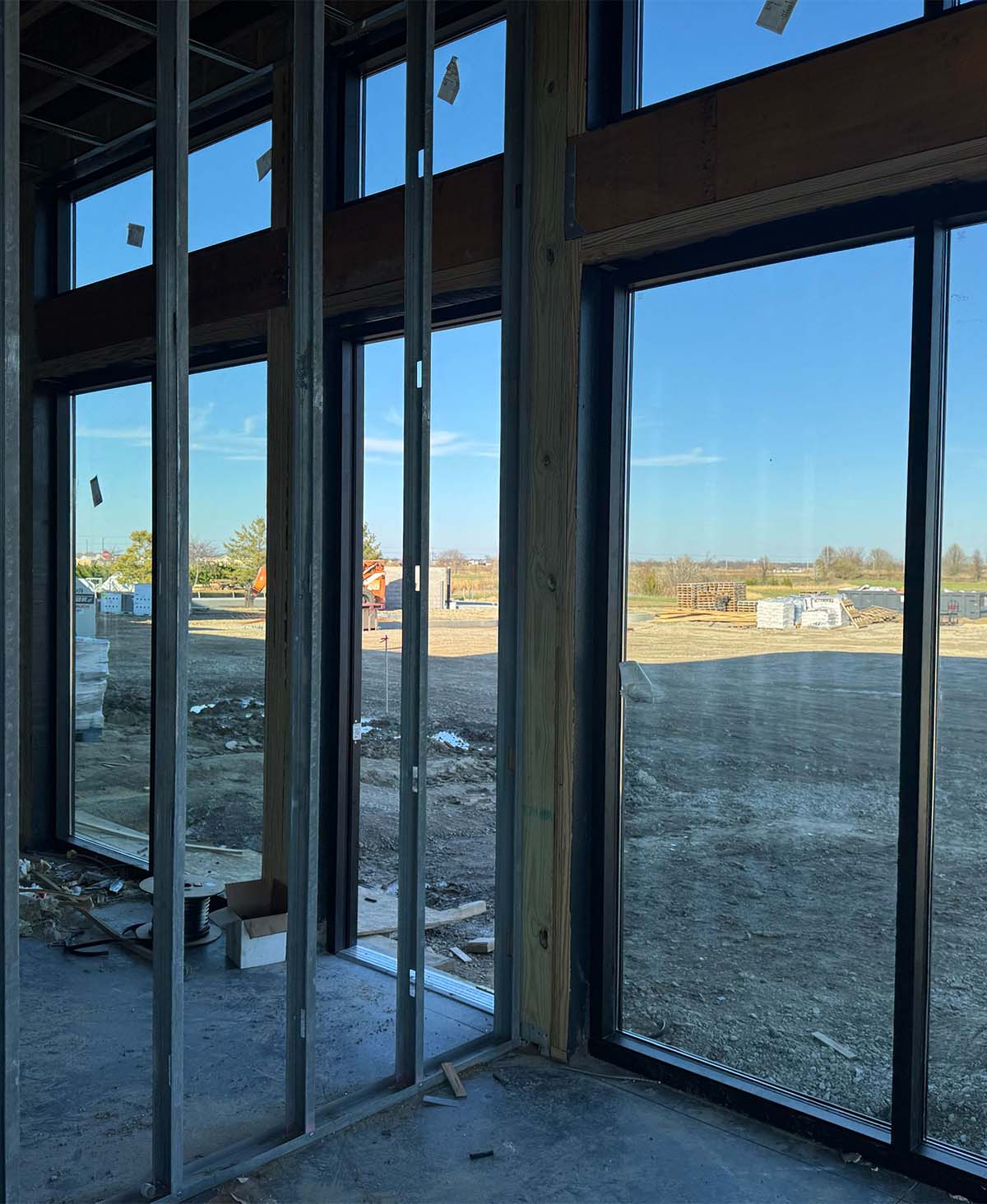 Interior of a building under construction with metal framing and large windows showing a dirt landscape.
