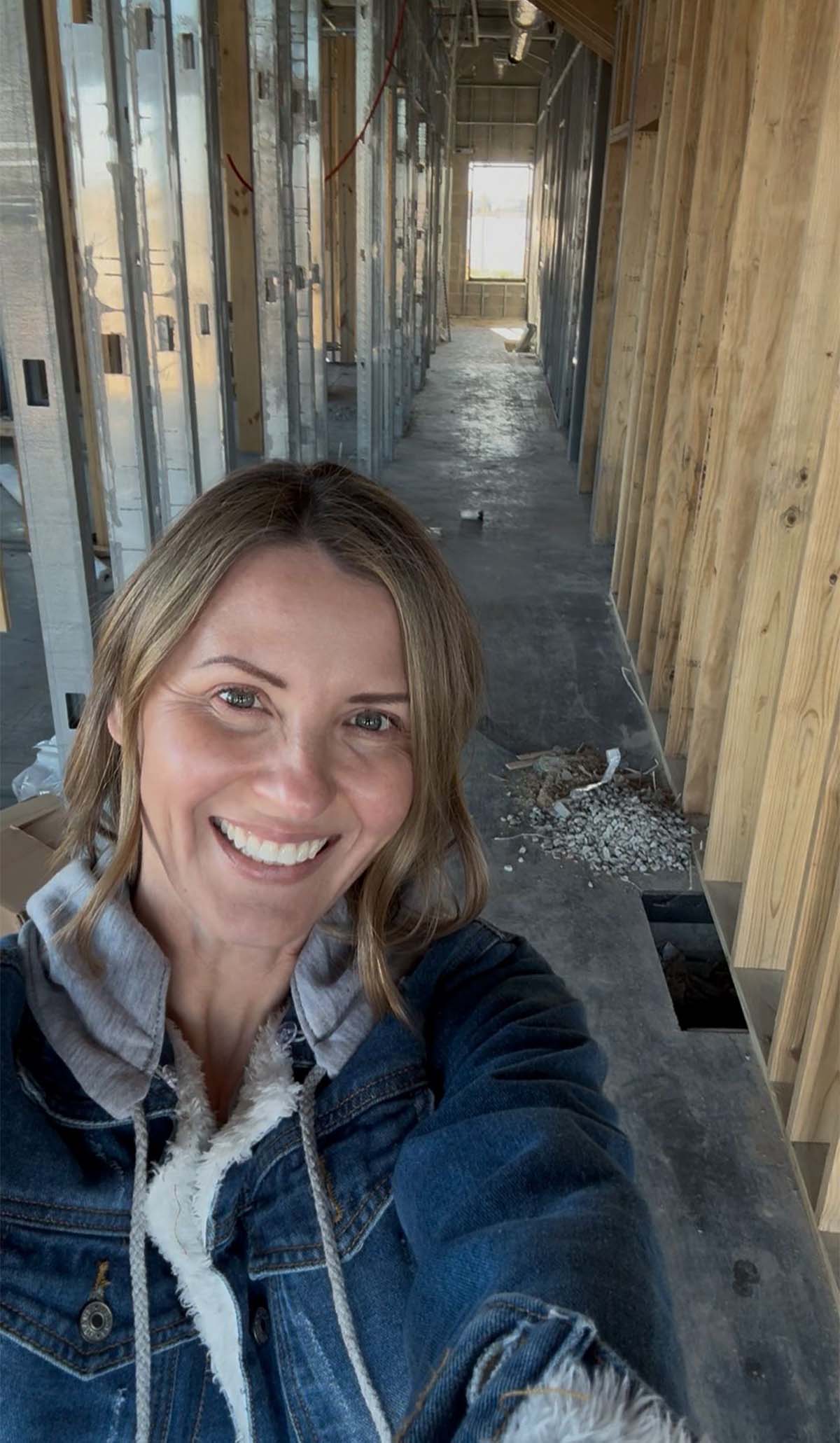 Smiling woman in a denim jacket taking a selfie inside a building under construction.