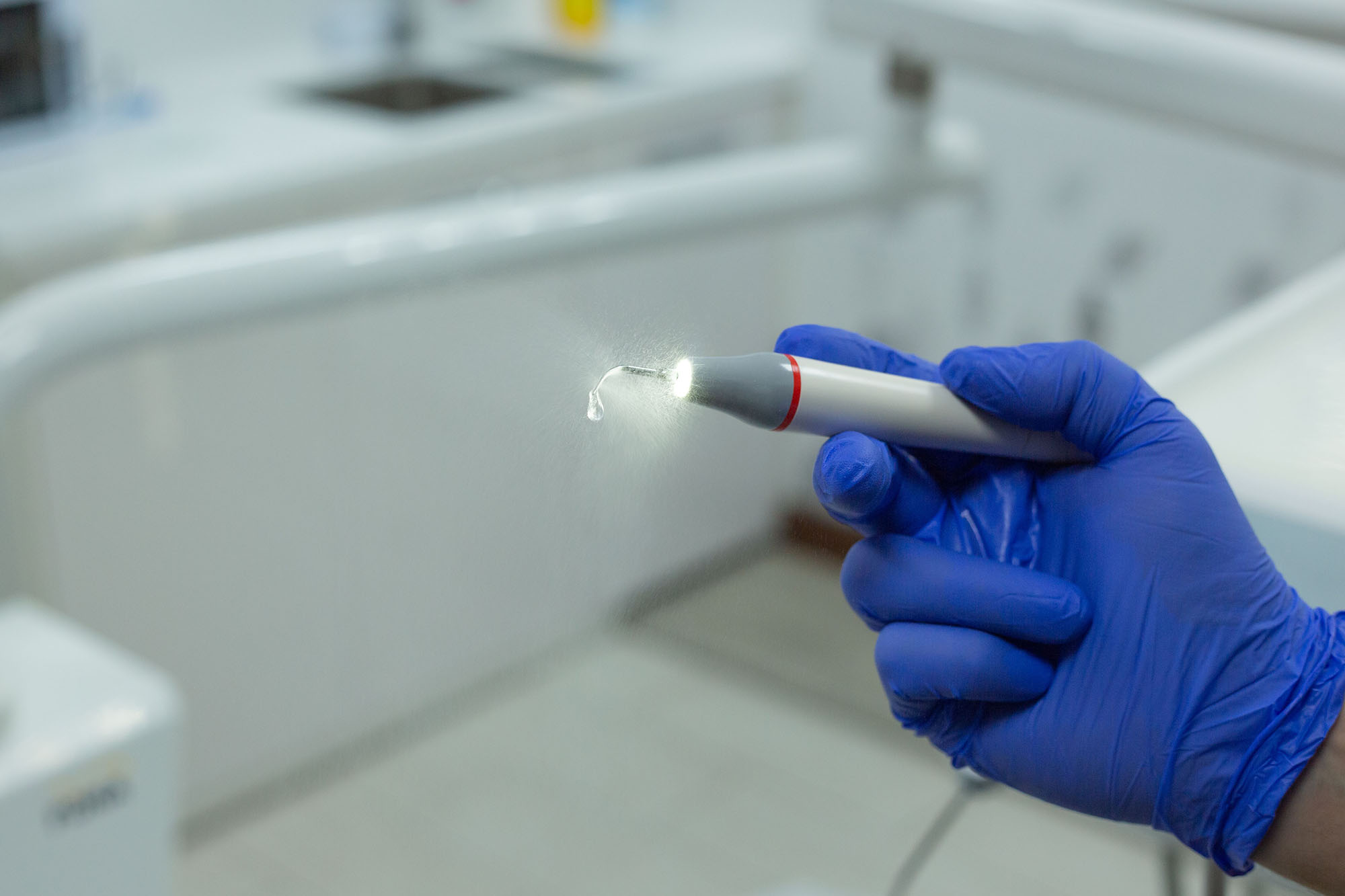Blue-gloved hand holding an illuminated ultrasonic scaler handpiece emitting a fine water mist in a dental office
