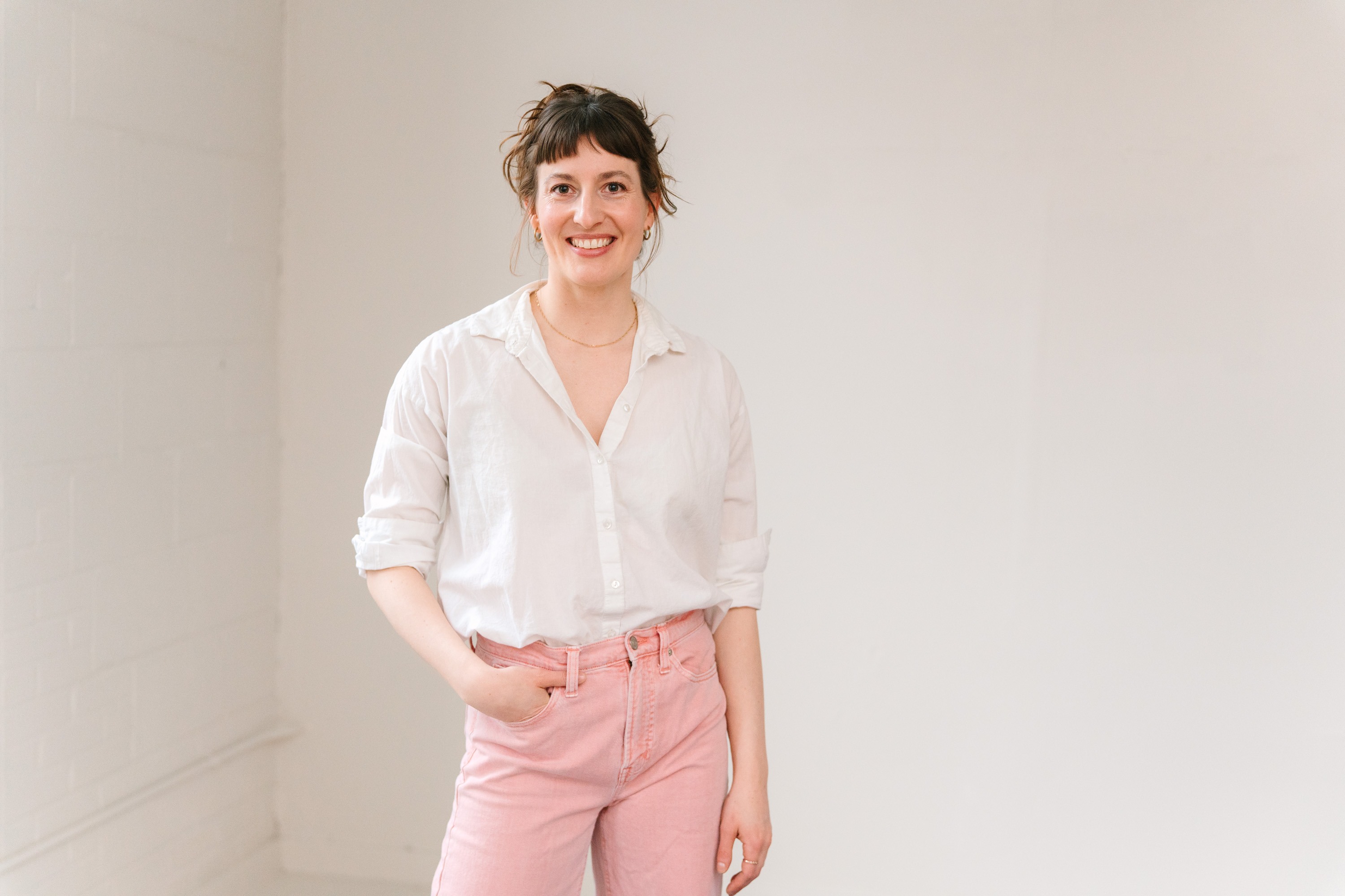 Smiling woman with brown hair in a casual updo wearing a white button-up shirt with rolled sleeves and pink pants, standing against a plain white wall with one hand in her pocket.