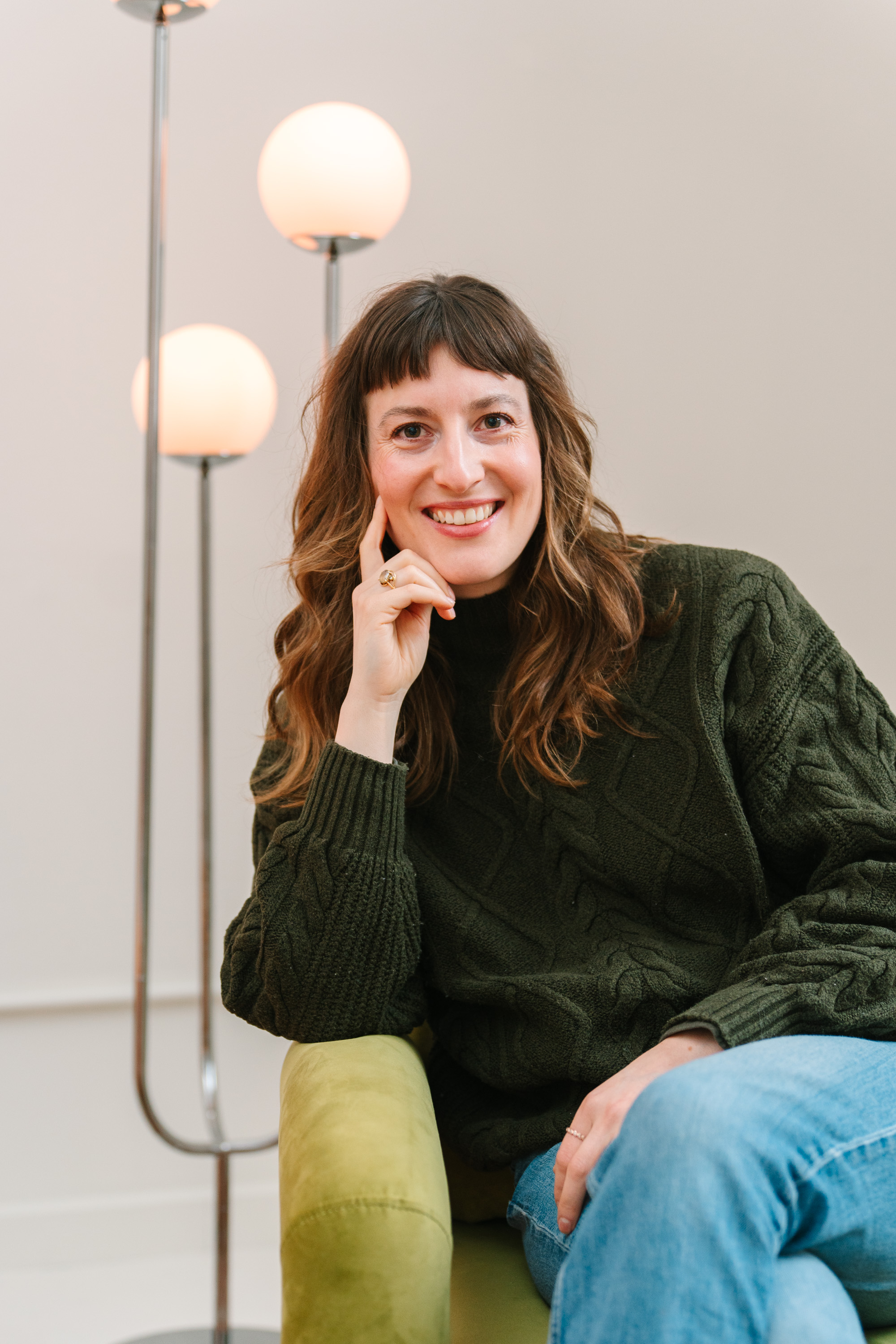 Smiling woman with shoulder-length brown hair and bangs sitting on a green chair, wearing a dark green knit sweater and blue jeans, with a floor lamp with three round lights in the background.