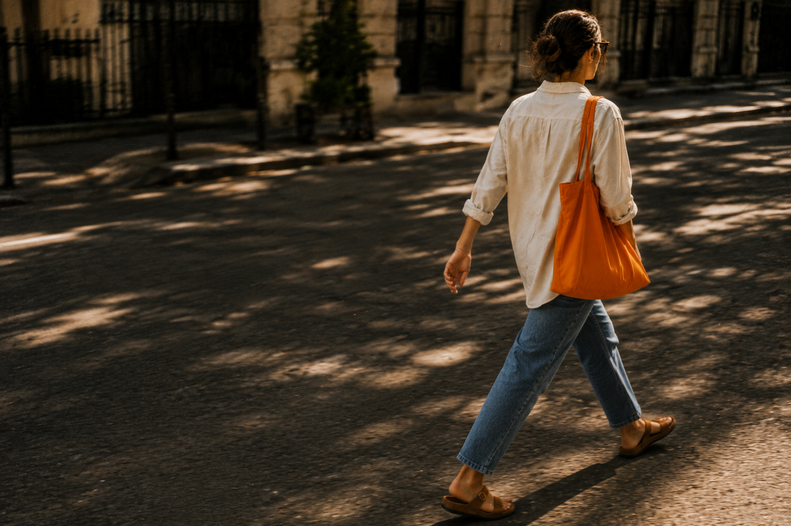 Woman walking down a sunlit street with an orange tote bag