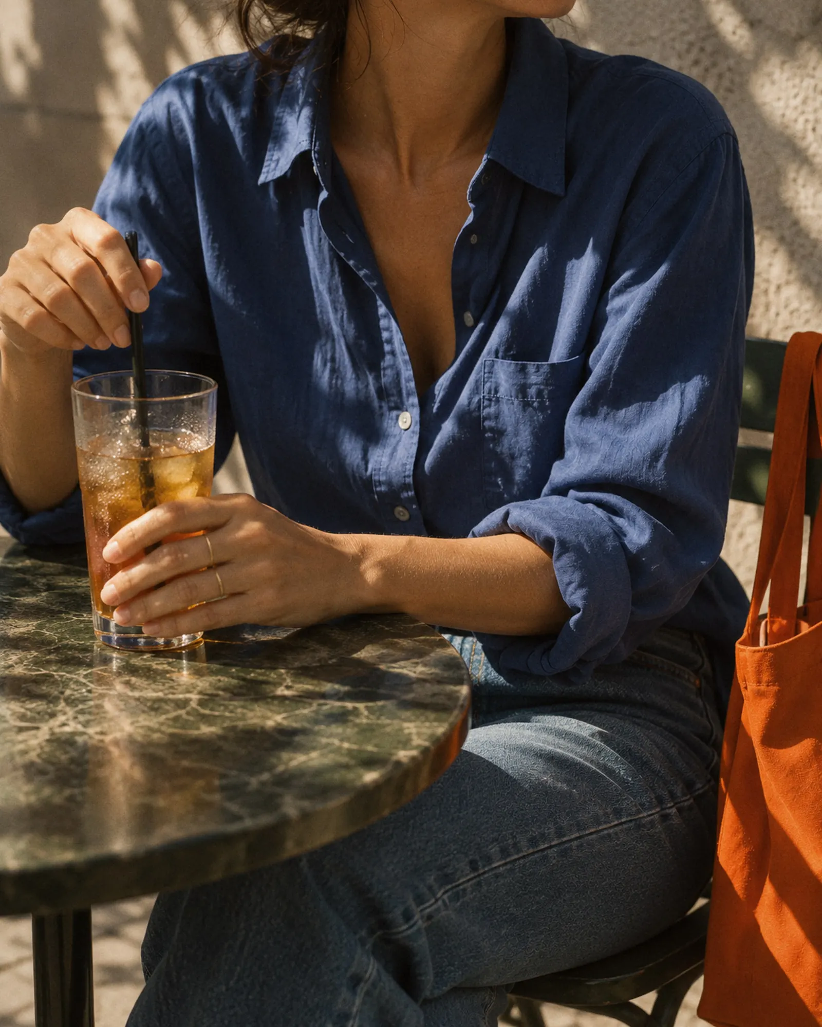 Woman in a blue linen shirt stirring an iced tea at a green marble cafe table