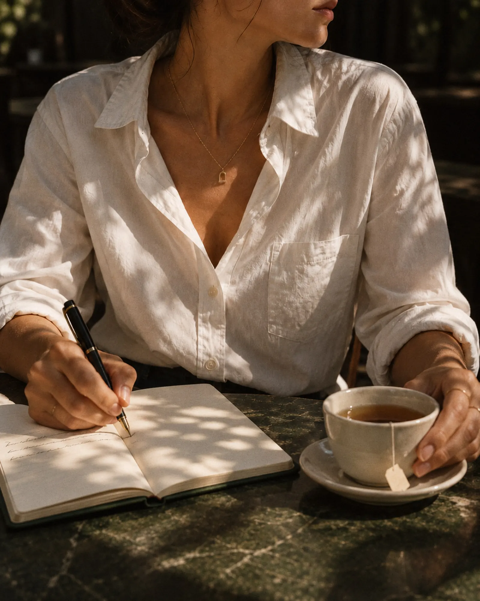 Hands writing in an open journal beside a cup of tea on a green marble cafe table