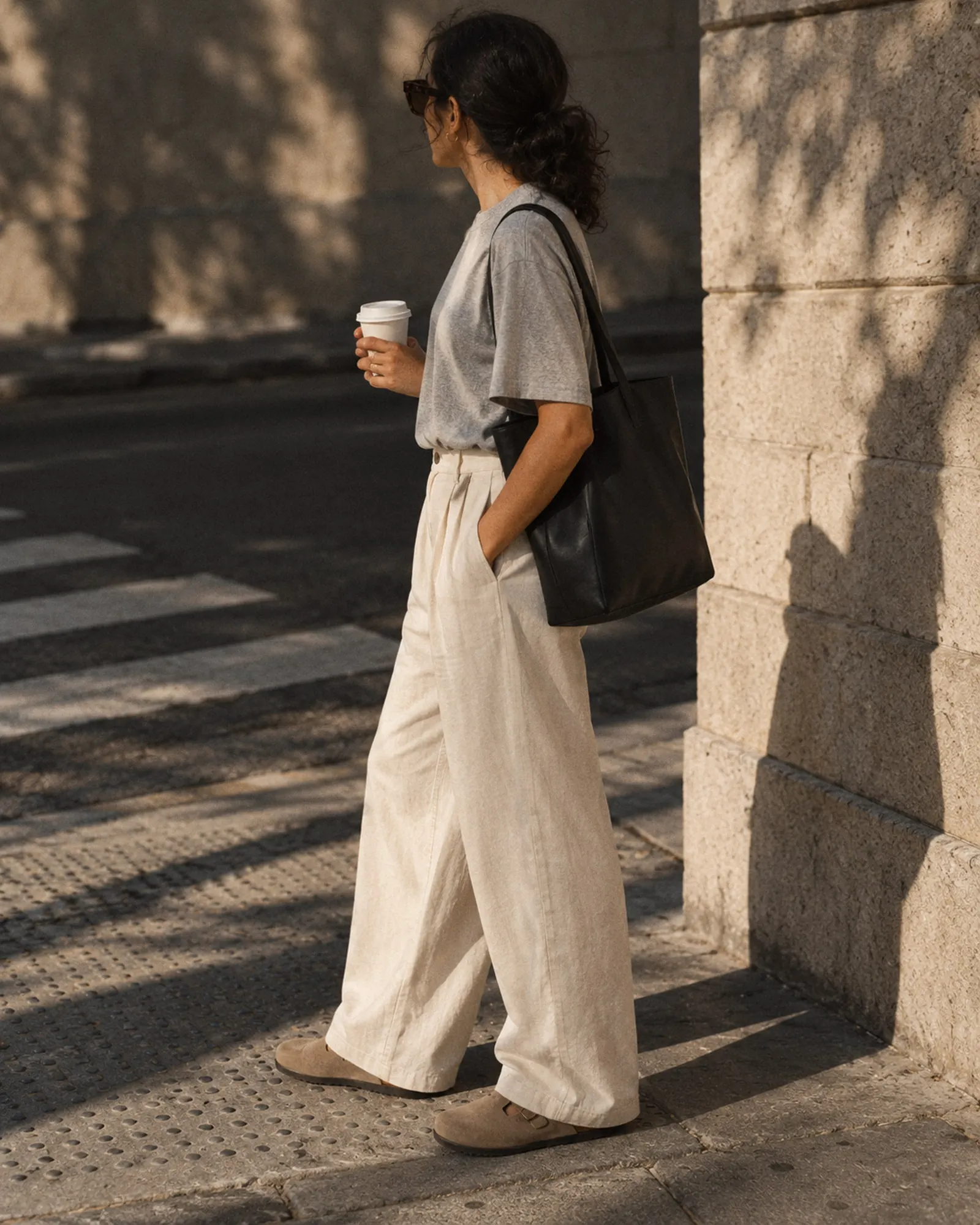 Woman in cream trousers and a grey tee standing on a street corner with a coffee and a black tote