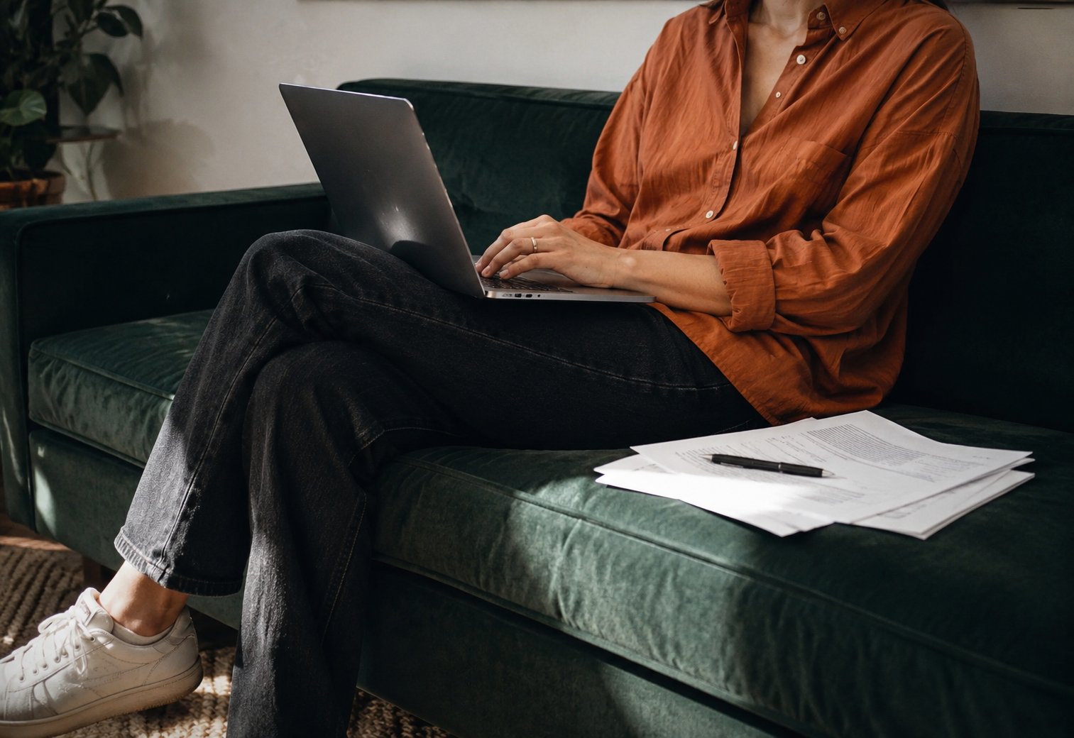 Woman in an orange shirt working on a laptop with papers beside her
