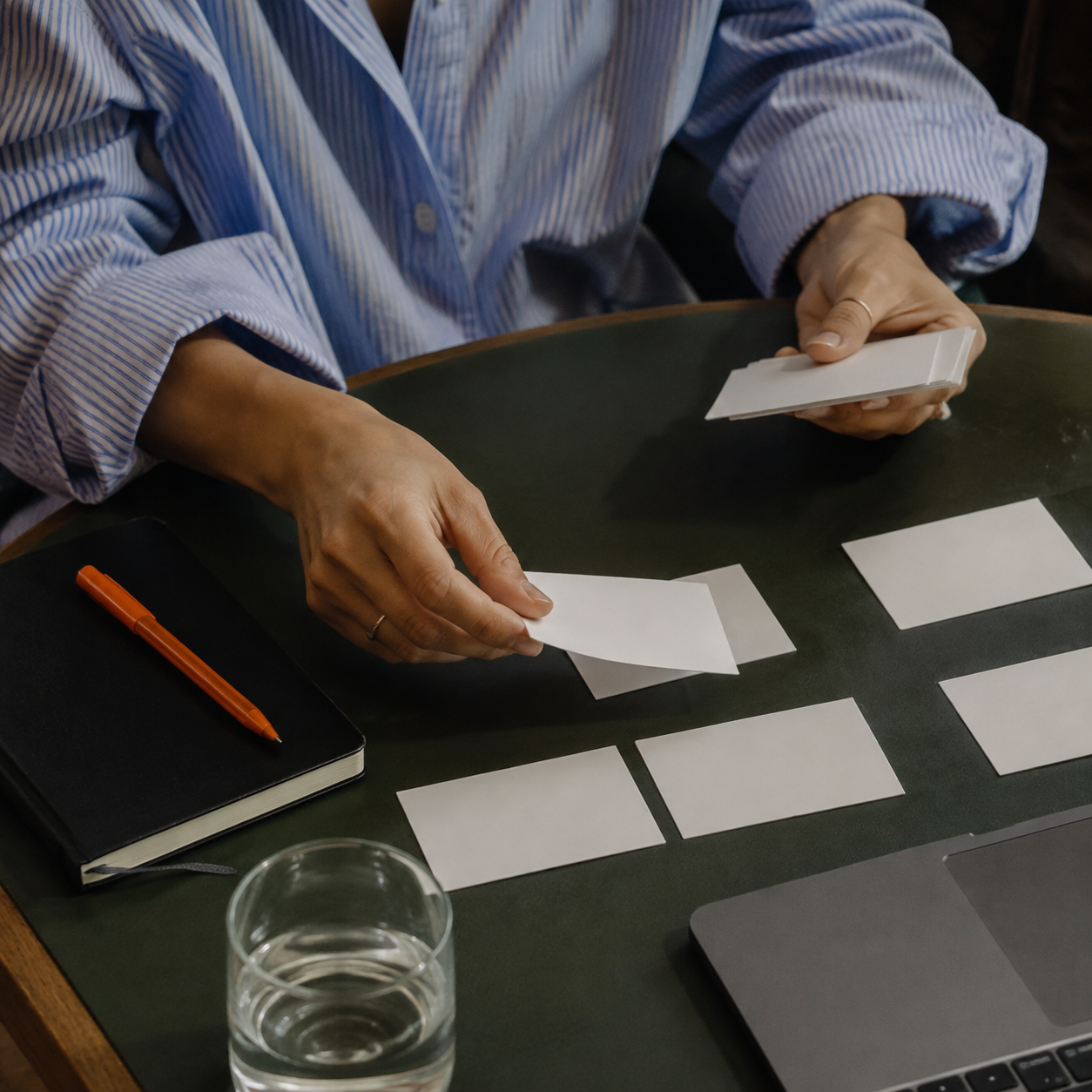 Hands sorting blank cards on a table beside a notebook and laptop