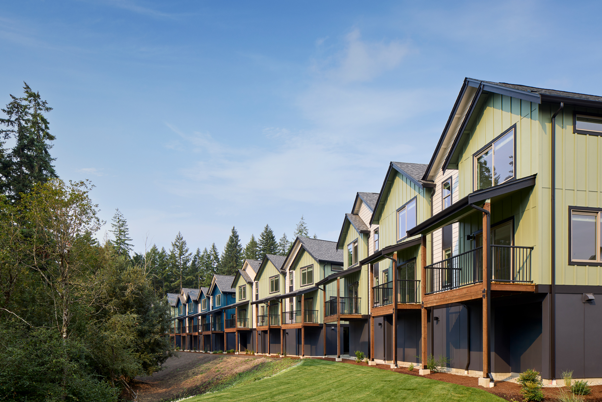 Row of modern two-story townhouses with elevated wooden balconies, painted in shades of green, blue, and cream, next to a grassy area and forest under a clear blue sky.
