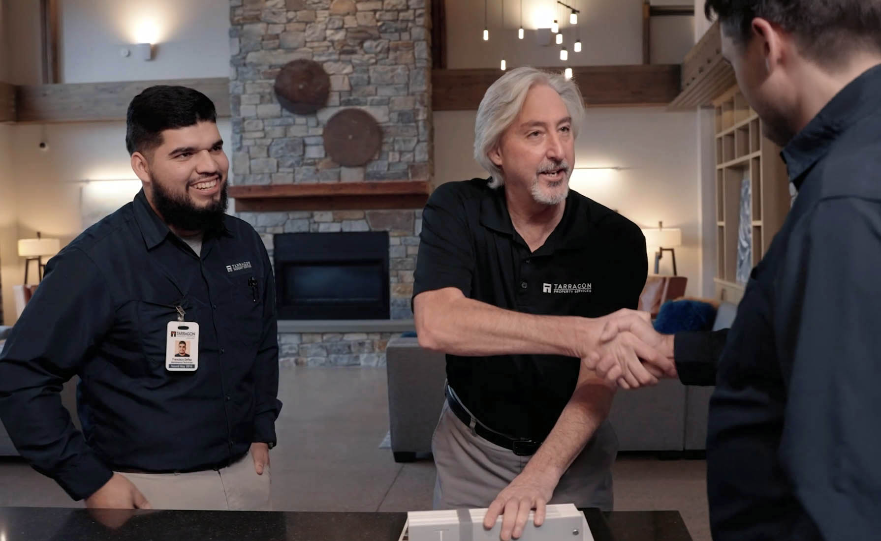 Three men in a modern living room; one man with gray hair wearing a black Tarragon Property Services polo shirt shakes hands with a man on the right, while another man with a beard in a dark shirt and beige pants smiles watching the handshake.