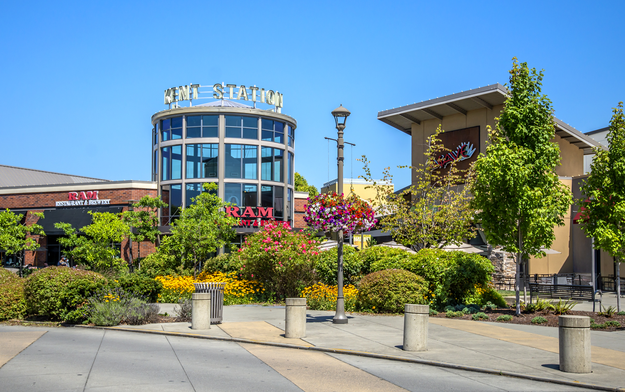 Exterior of Kent Station shopping area featuring a round glass building with 'Kent Station' sign, surrounded by lush greenery, colorful flowers, and adjacent store signs including RAM Restaurant & Brewery under clear blue sky.