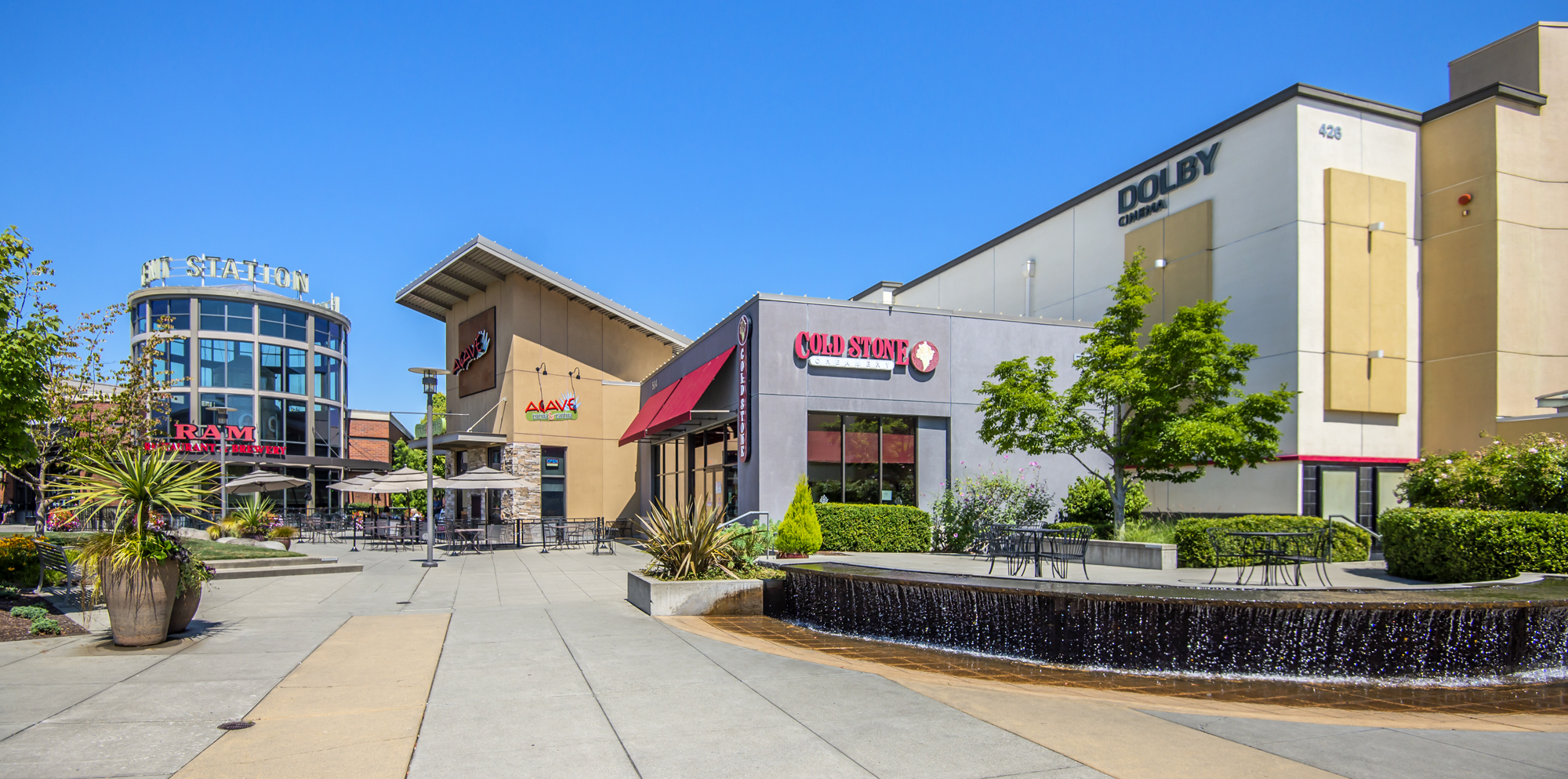 Outdoor plaza with patio seating, large potted plants, and a water feature in front of commercial buildings housing Cold Stone Creamery, Dolby Cinema, and Agave Mexican Kitchen, under a clear blue sky.