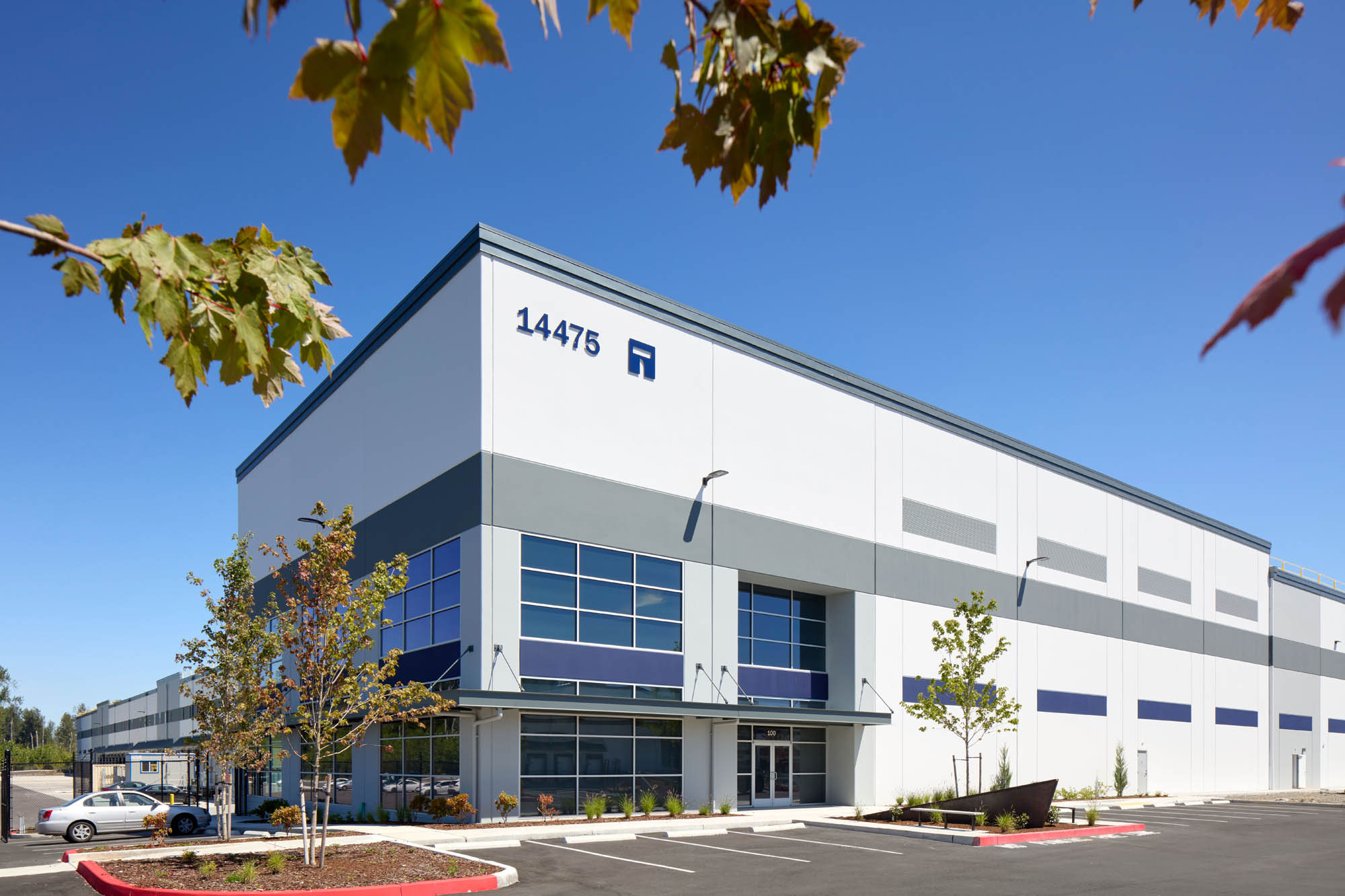 Modern industrial warehouse building with large windows, gray and white exterior, marked with number 14475, surrounded by young trees and an empty parking lot under a clear blue sky.