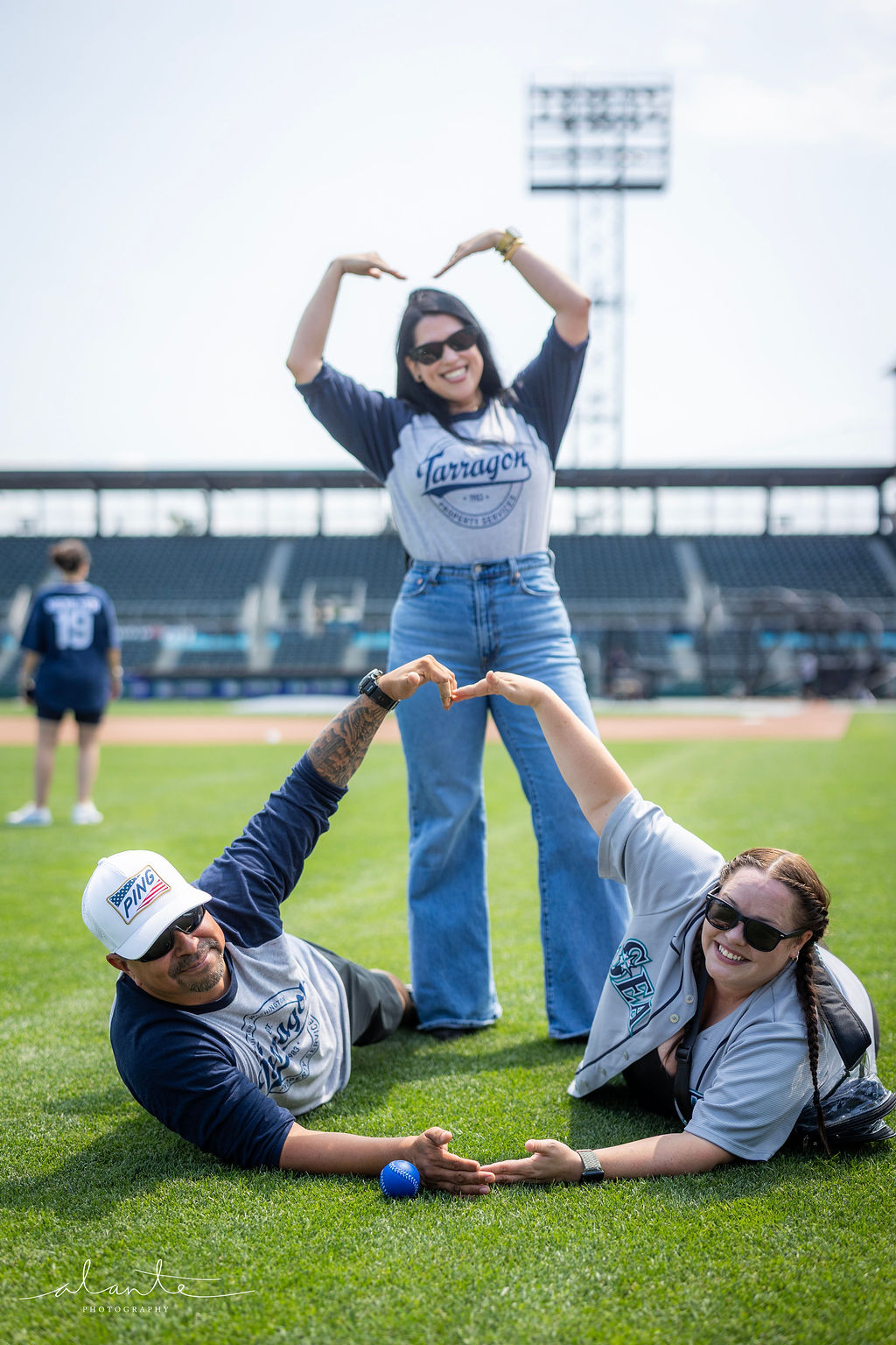 Three people on a baseball field making heart shapes with their arms and hands; one person stands smiling with sunglasses and a Tarragon shirt while two others lie on the grass forming the bottom of the heart with their arms, one wearing a white PING cap and the other in a gray shirt with a ponytail and sunglasses.