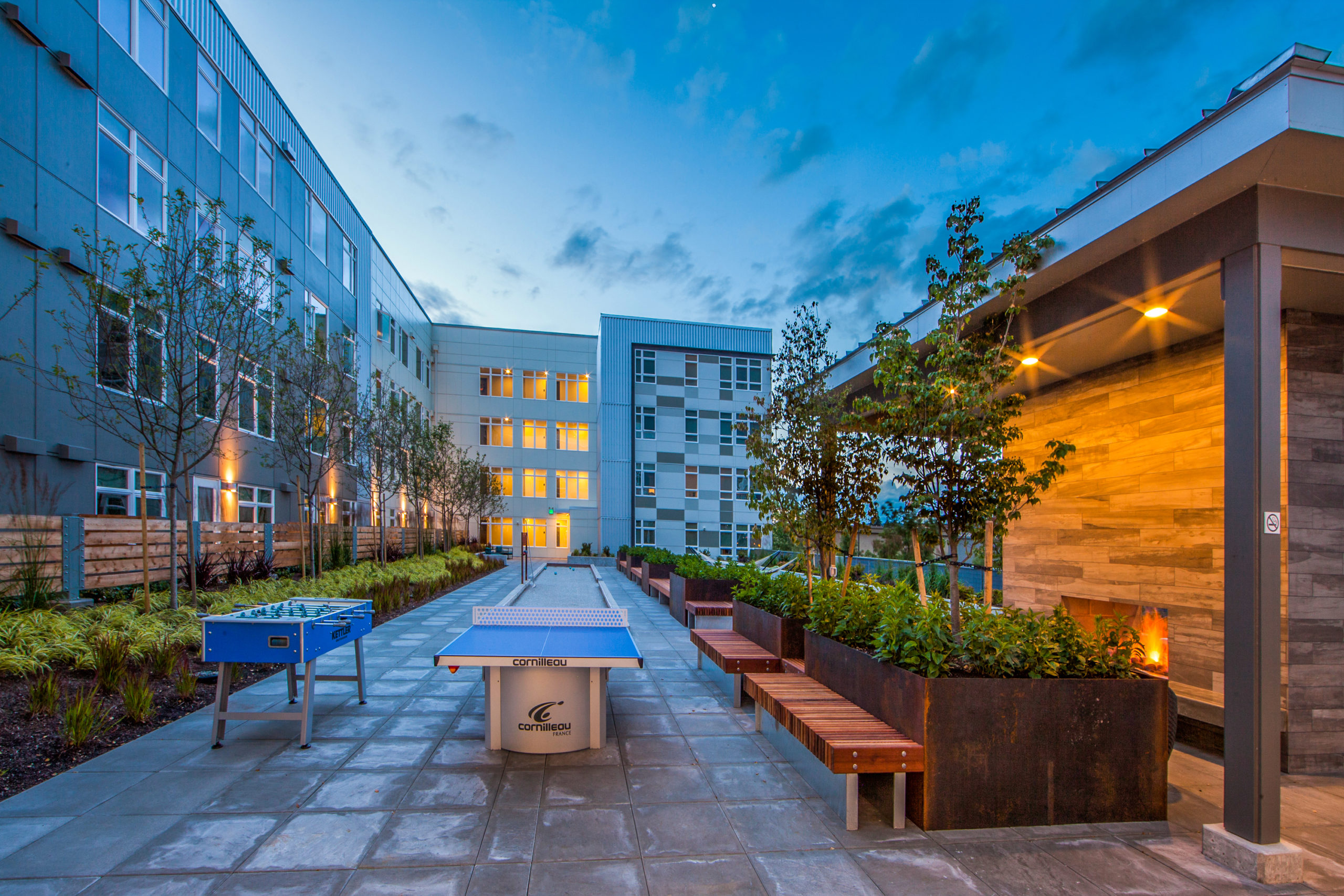 Outdoor courtyard of a modern apartment complex at dusk with a ping pong table, foosball table, bocce ball court, wooden benches with planters, and a covered fireplace area with glowing lights.