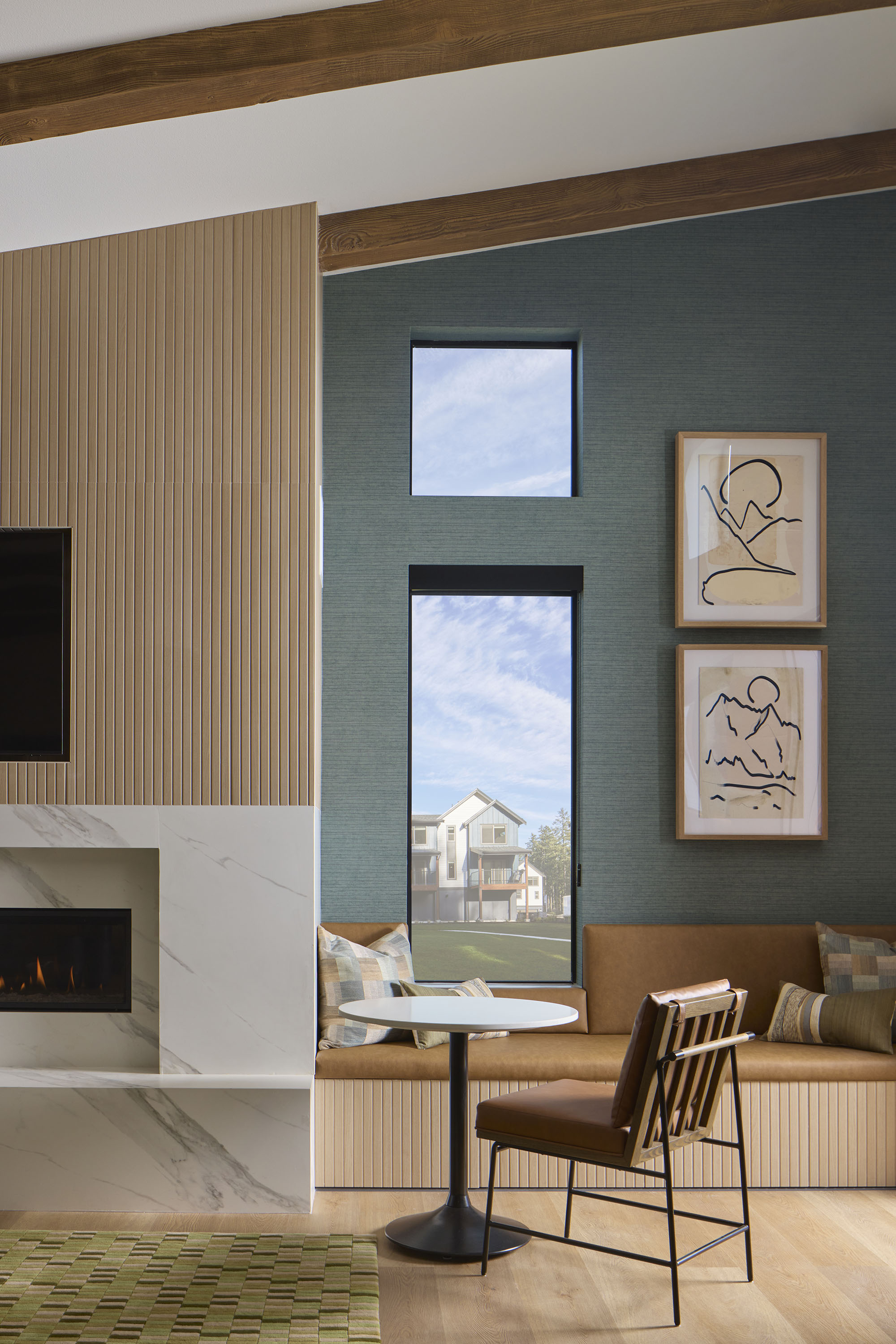 Modern living room corner with a built-in fireplace with marble surround, a brown leather cushioned bench with plaid and striped pillows, a round white table, and a brown cushioned metal chair, set against a blue textured wall with two abstract framed artworks and two vertically aligned windows showing a house and trees outside.