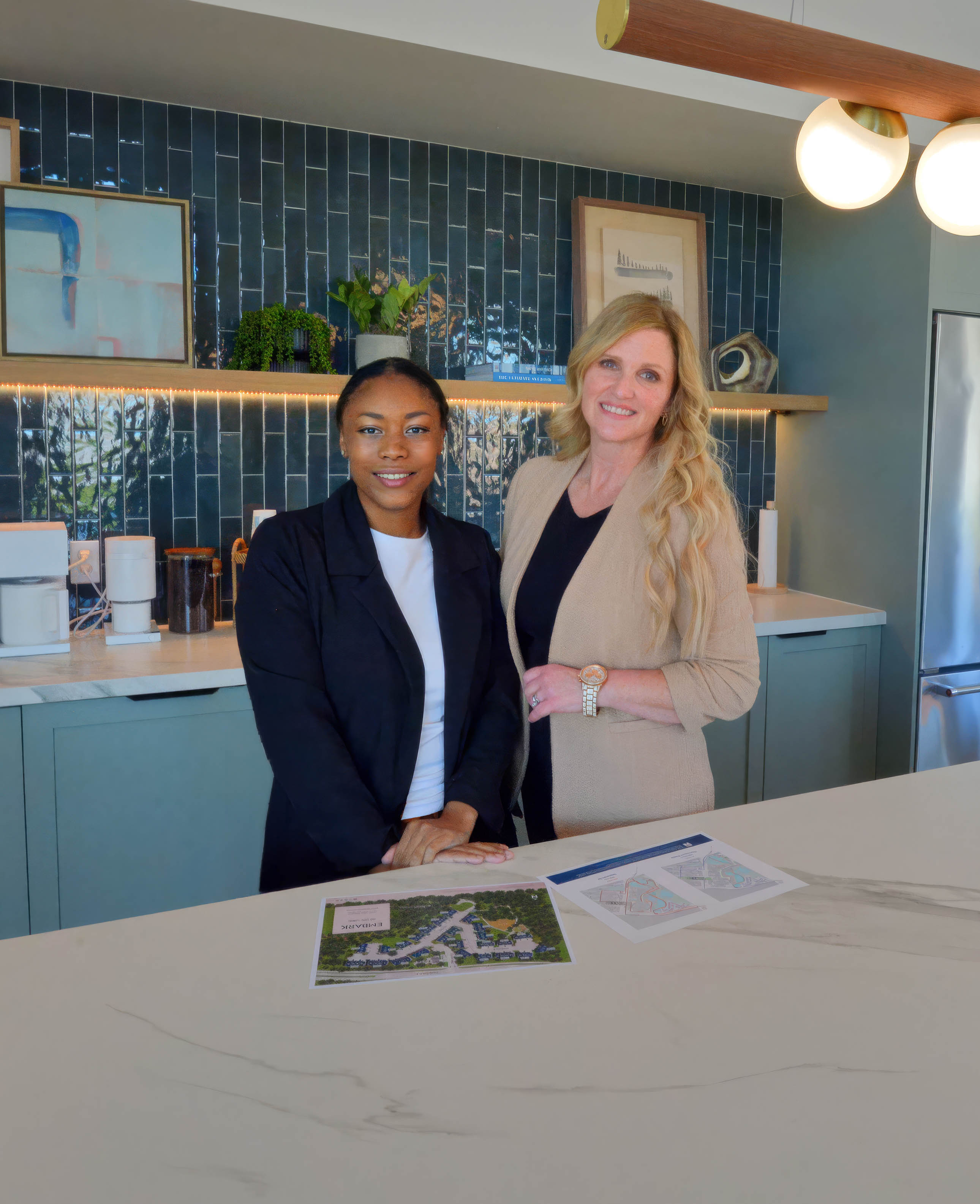 Two women standing behind a kitchen counter with architectural plans on it, smiling at the camera, with dark tiled wall and shelves with plants and framed art in the background.