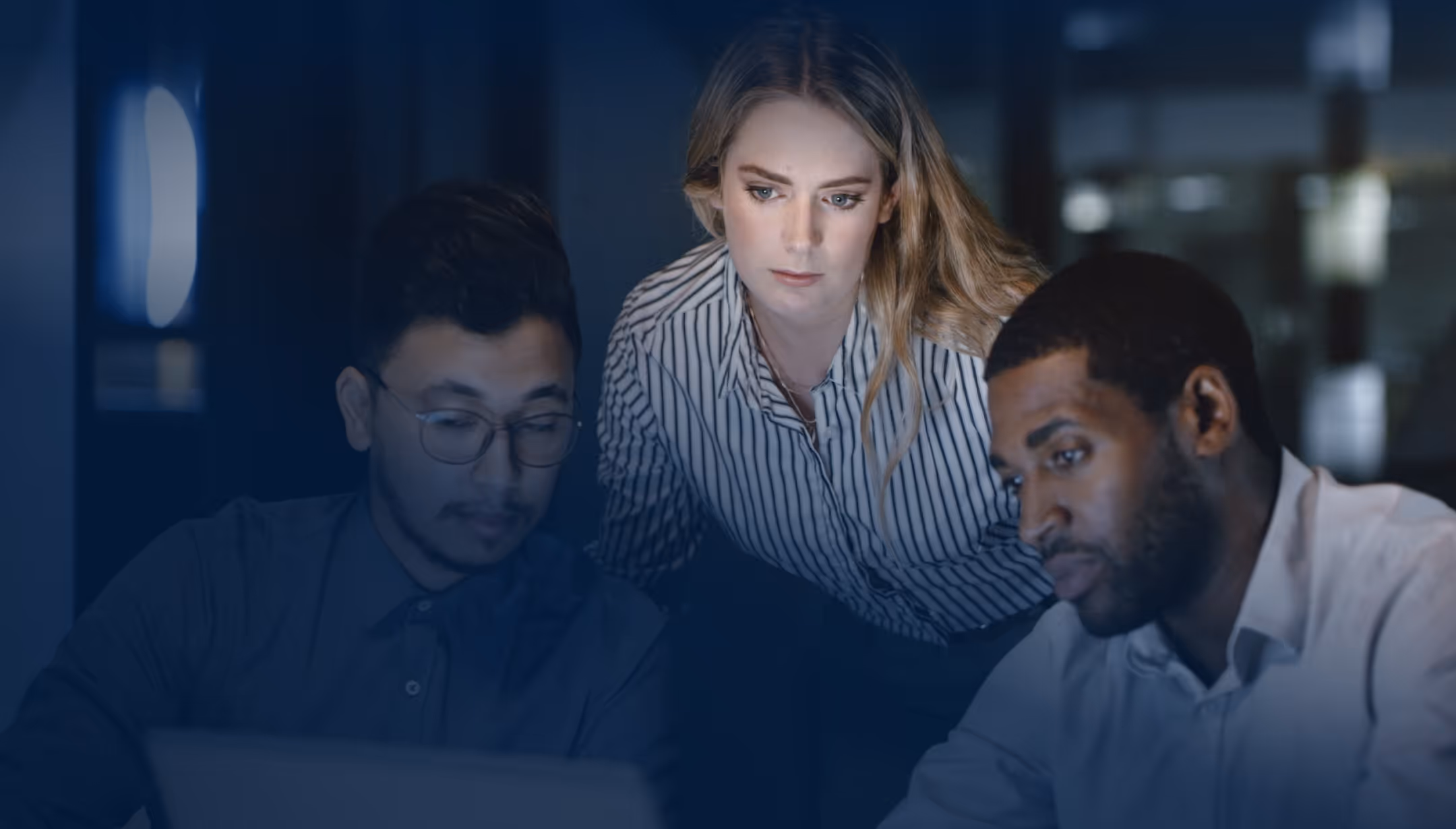 Three diverse coworkers focused on a laptop during a nighttime work session in an office.