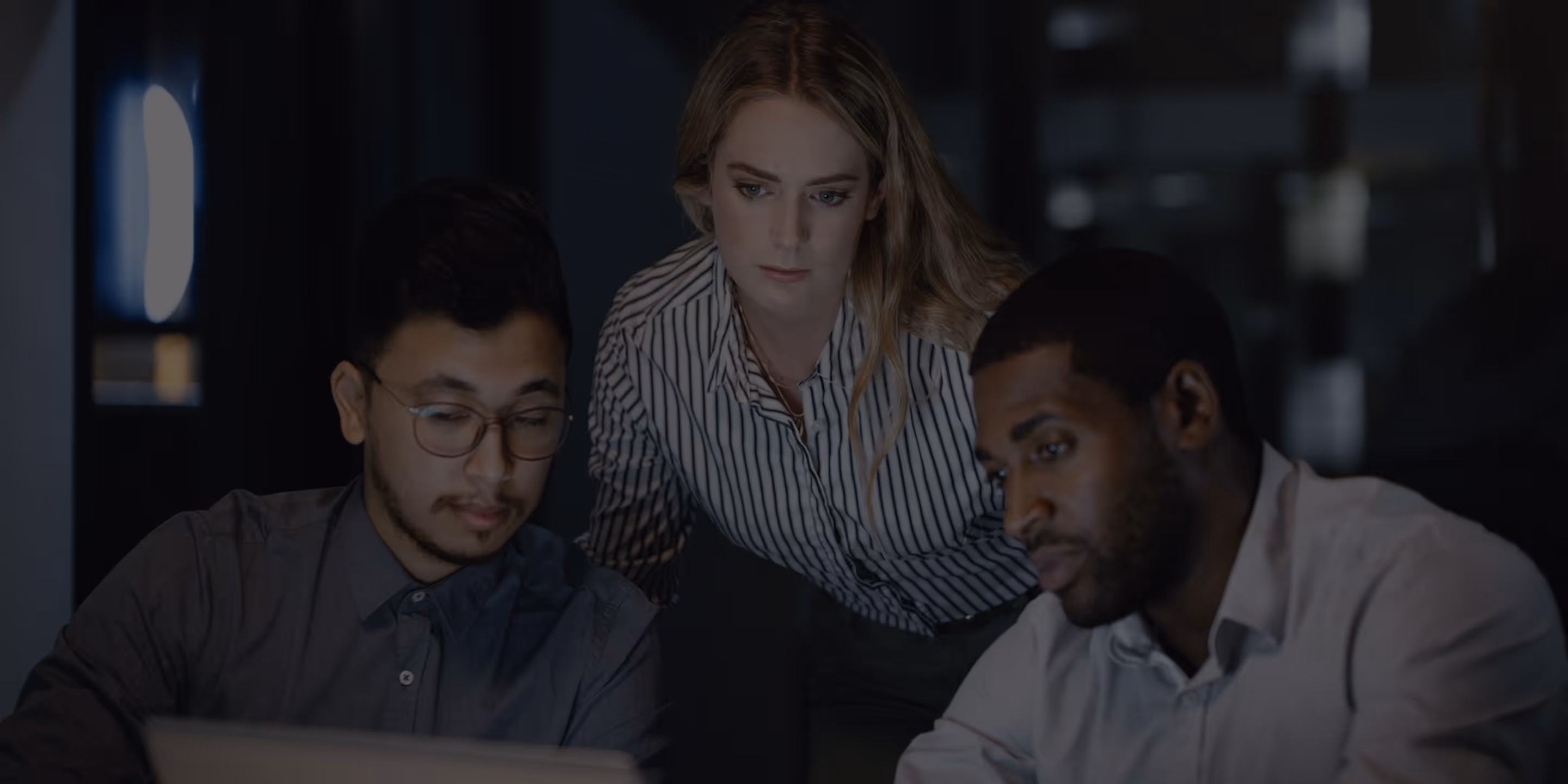Three coworkers focused on a laptop screen in a dimly lit office late at night.