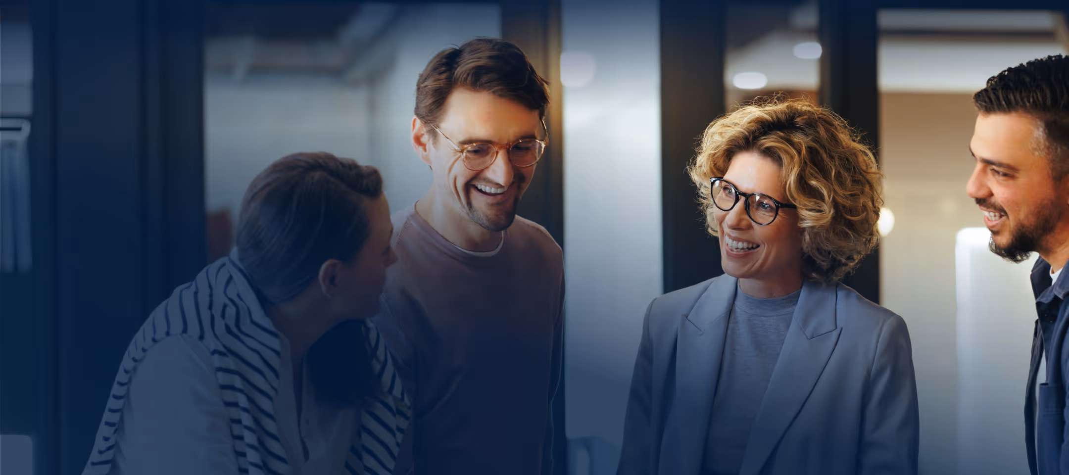 Four diverse coworkers smiling and talking together in a modern office setting.