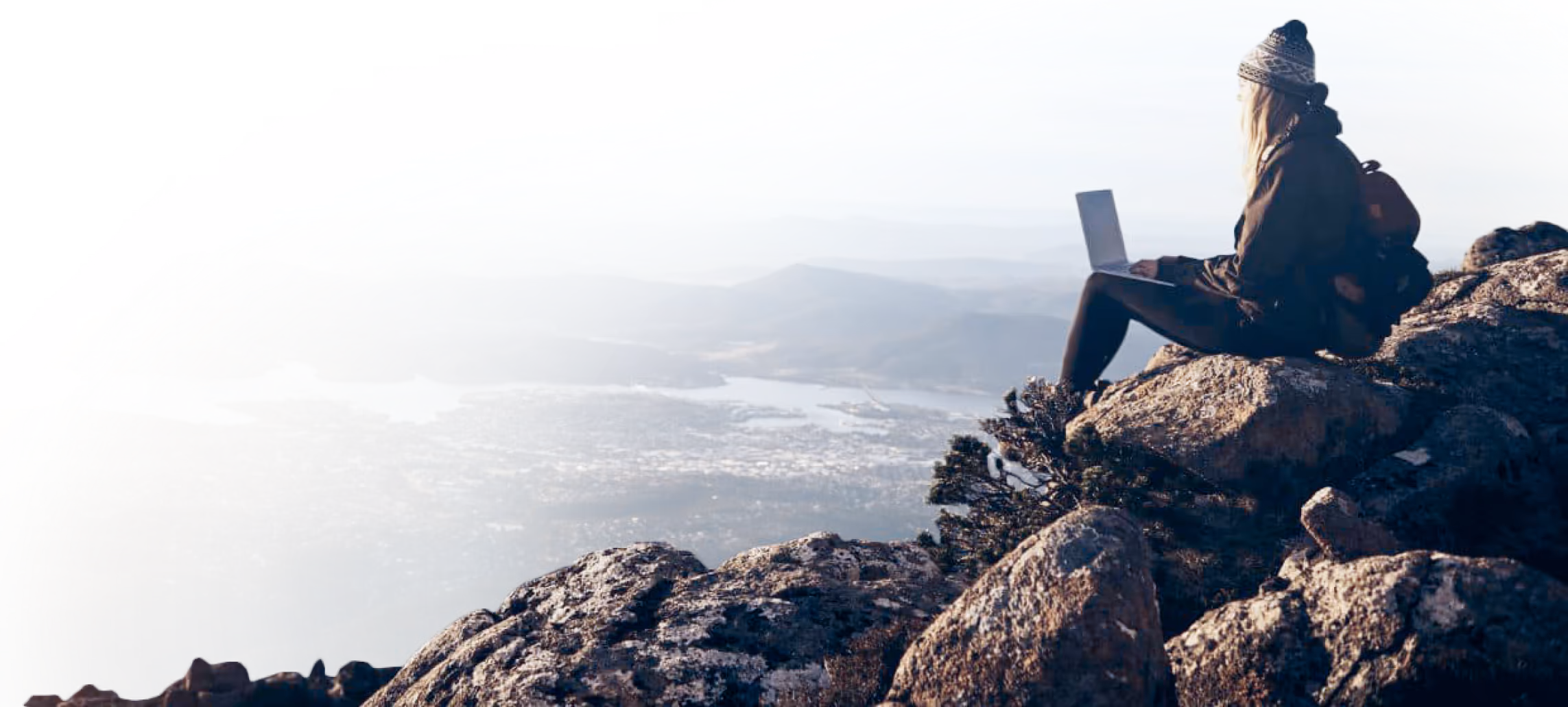 Person sitting on rocky mountain peak using laptop with a scenic valley and lake view below.