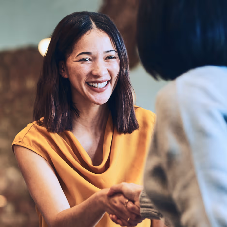 Smiling woman in a yellow top shaking hands with another person in a gray sweater.