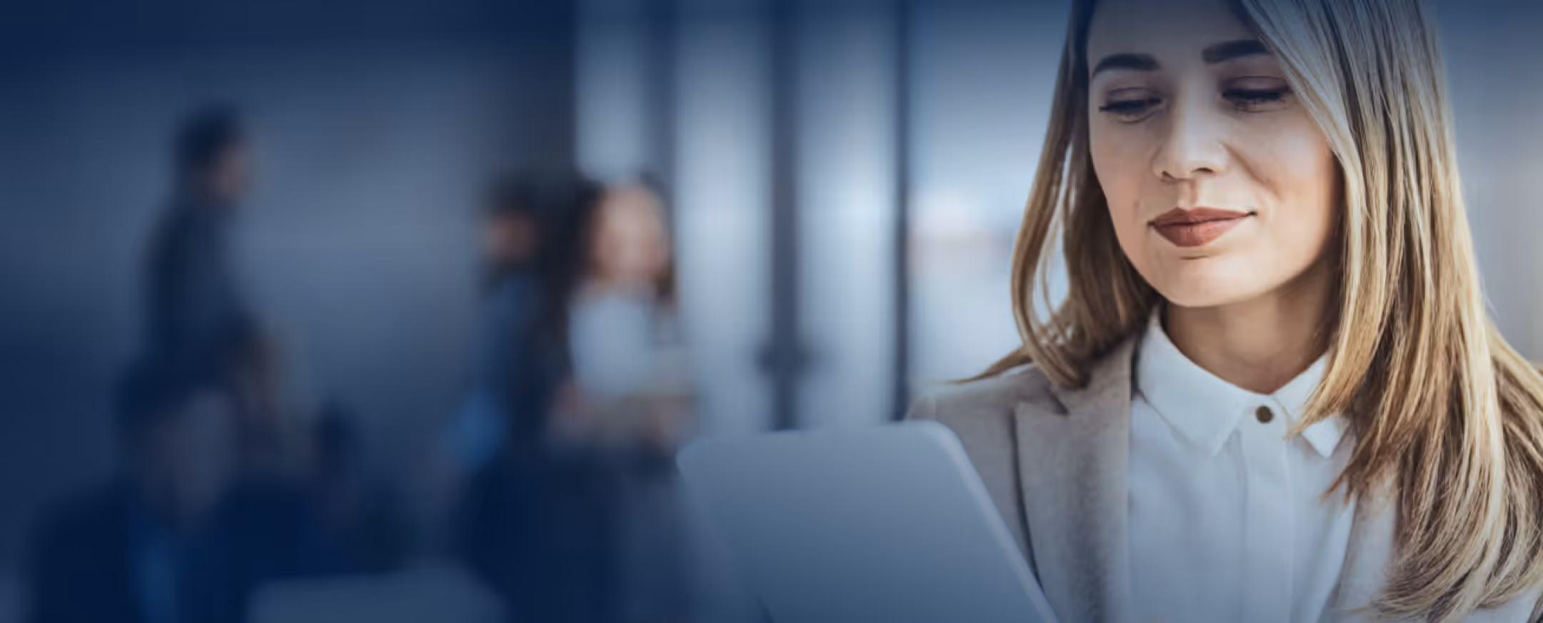 Smiling woman in blazer looking at a tablet with blurred people in a modern office background.