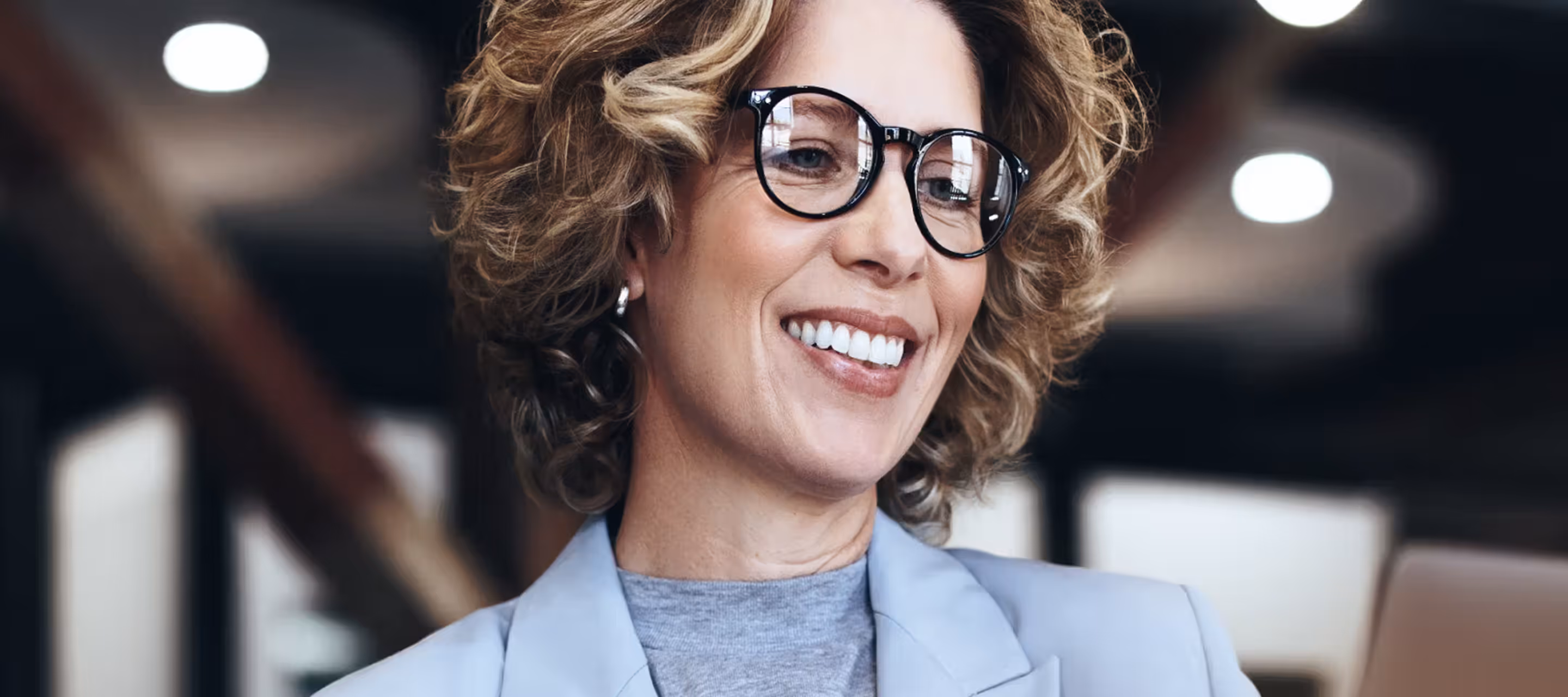 Smiling woman with curly hair and glasses wearing a light blazer in an office setting.