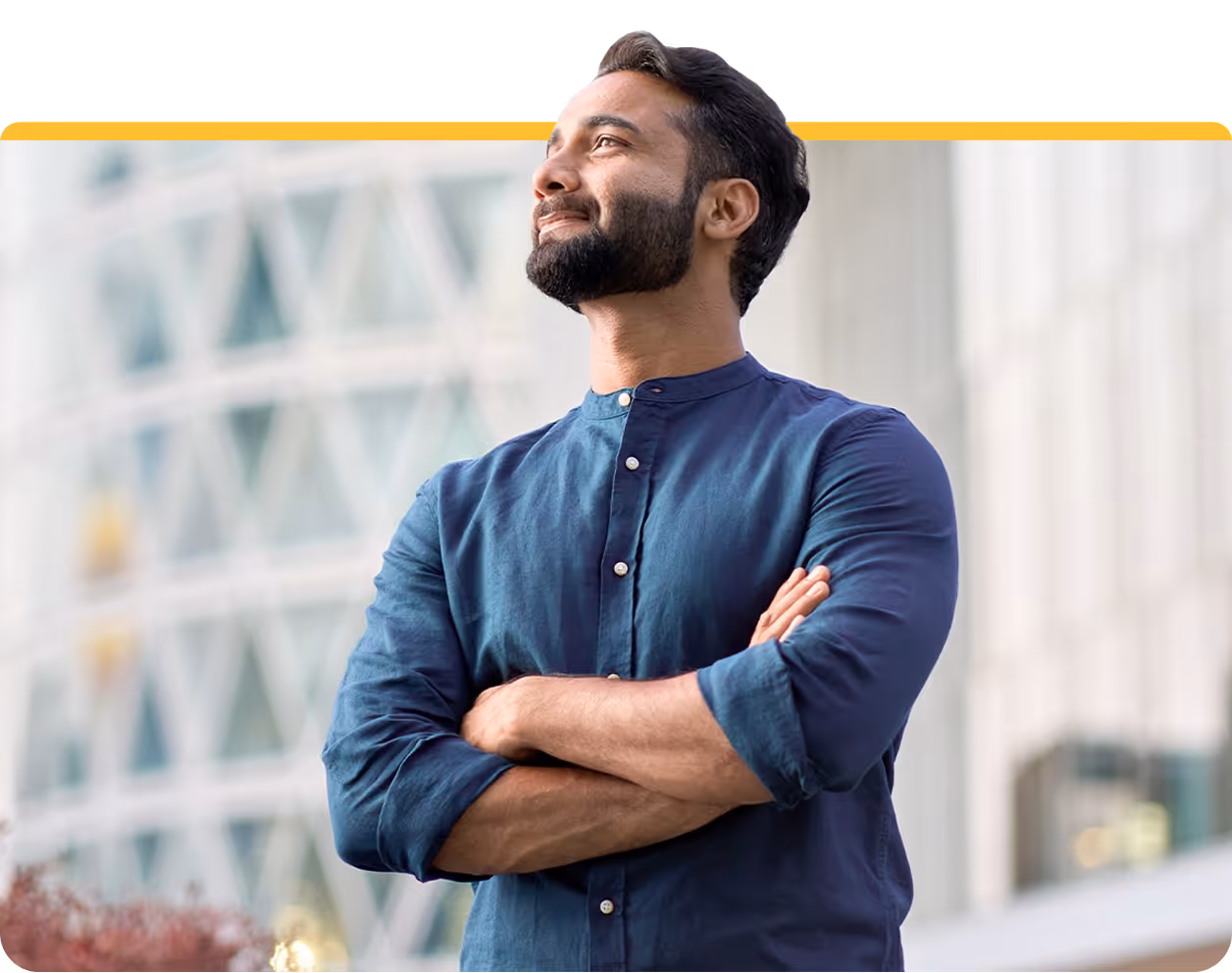 Bearded man in blue shirt with arms crossed looking upward thoughtfully outdoors in urban setting.