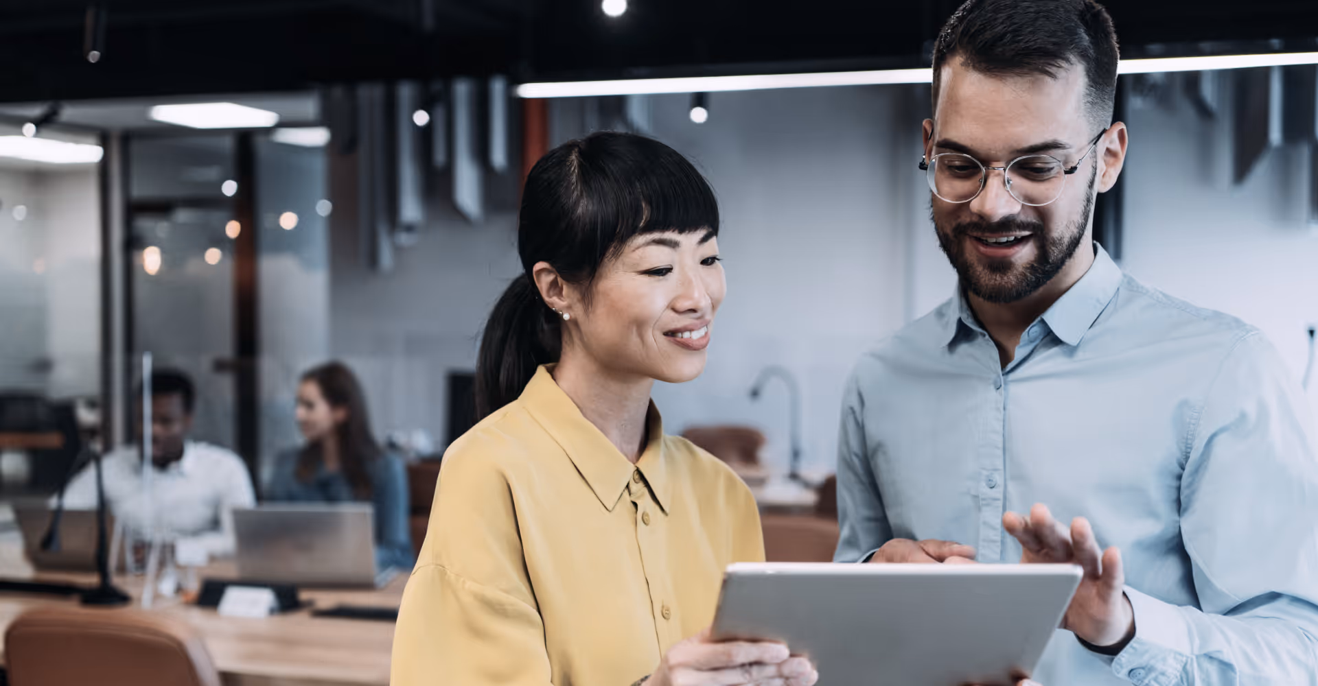 Two colleagues, a woman and a man, smiling and looking at a tablet in a modern office.