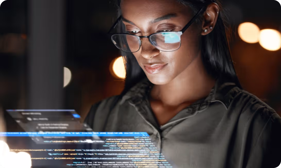 Woman wearing glasses looking at glowing computer code on a transparent screen in low light.