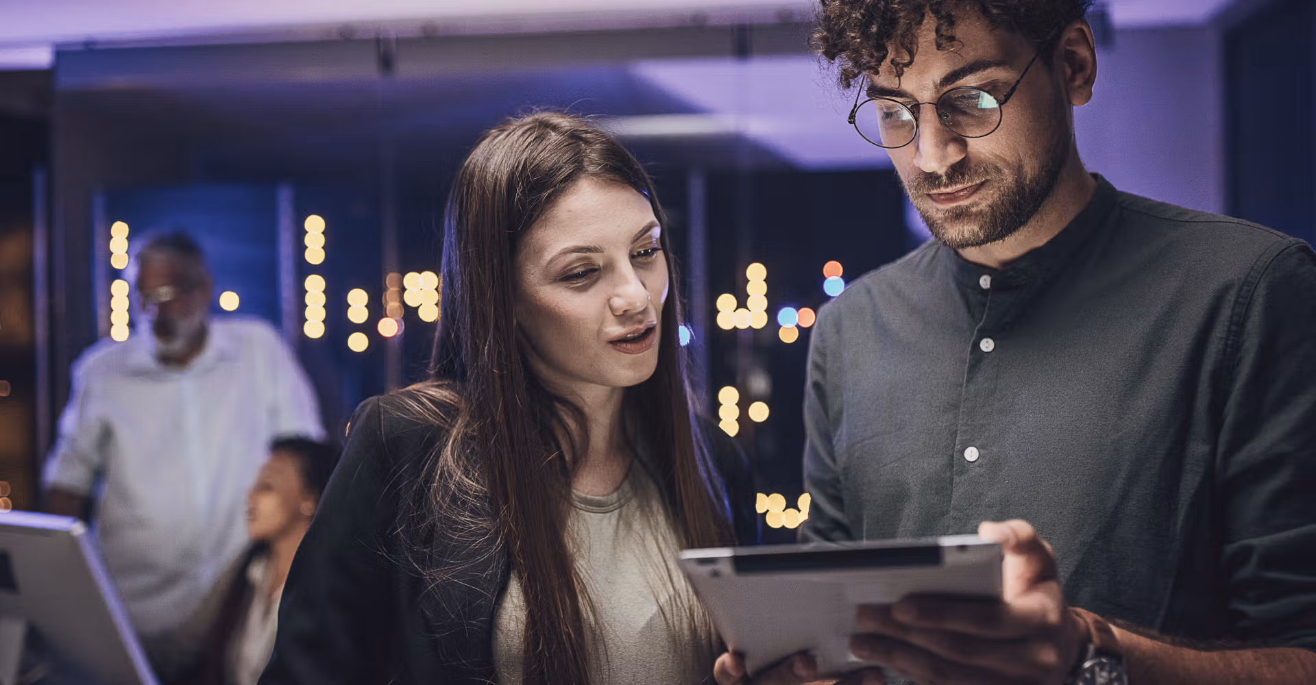 Two coworkers review a tablet while two other people work on computers in a modern office setting.