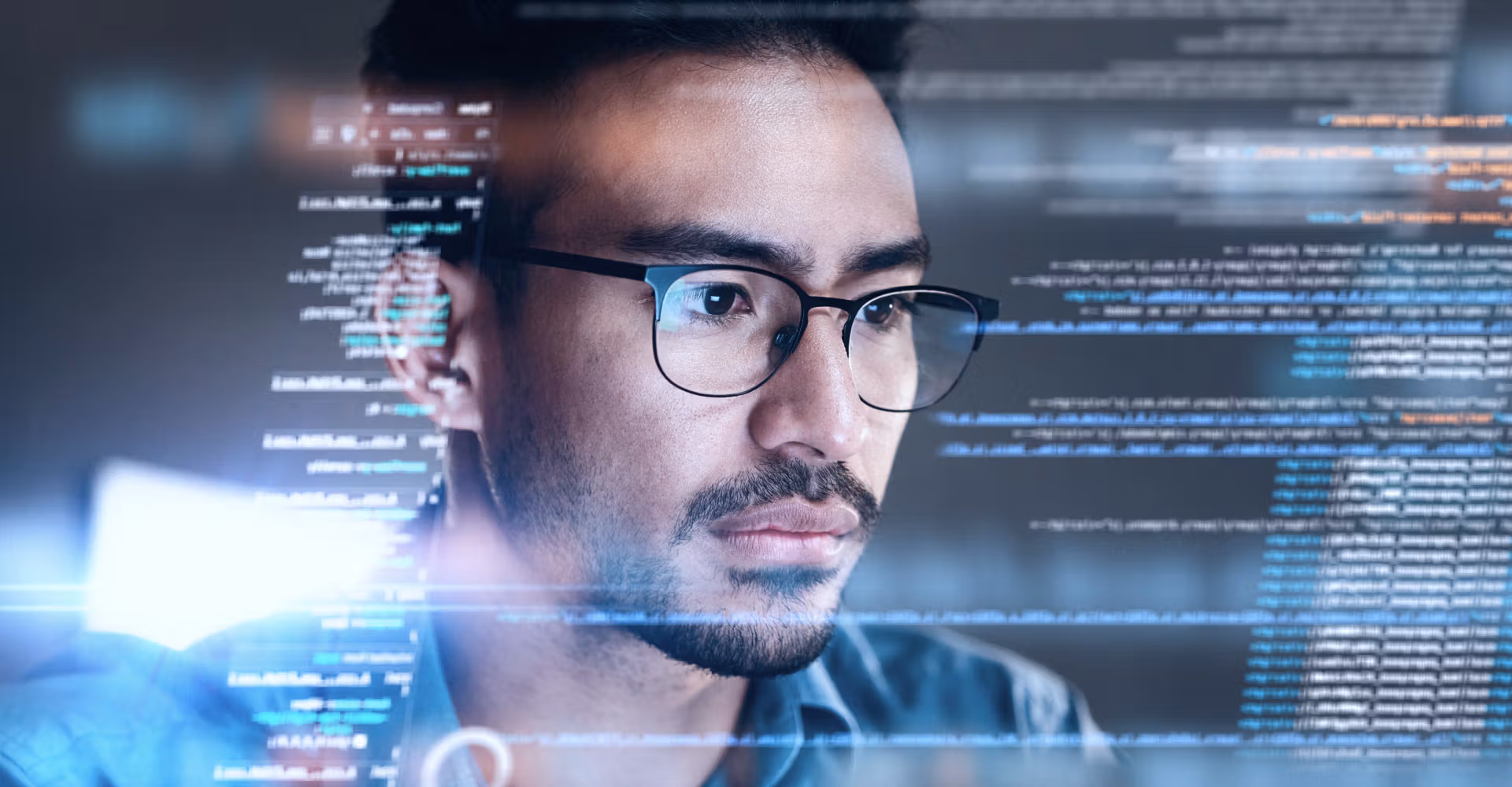 Man with glasses focused on computer screen with lines of code overlay.
