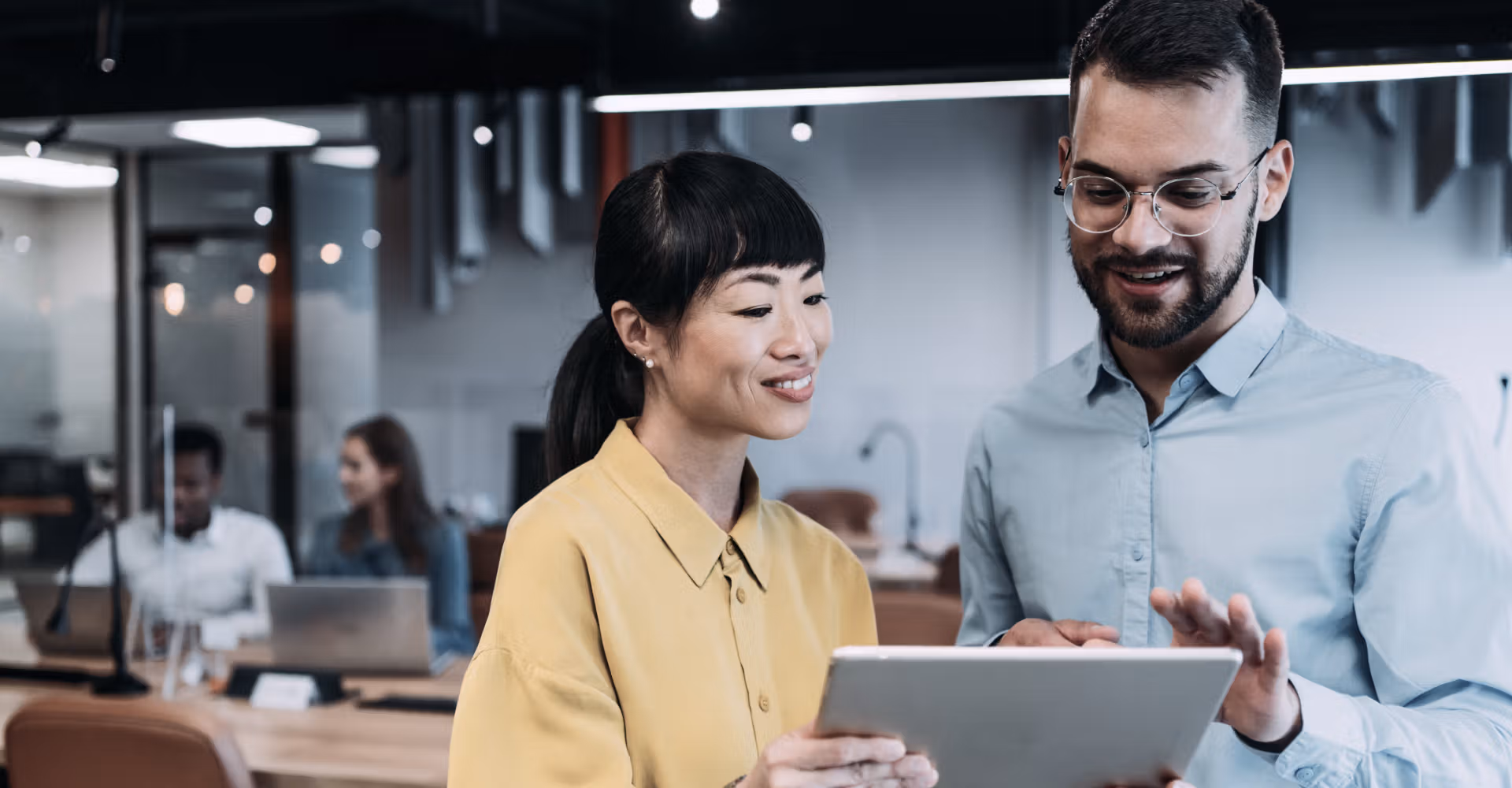 Two coworkers smiling and looking at a tablet together in a modern office setting.