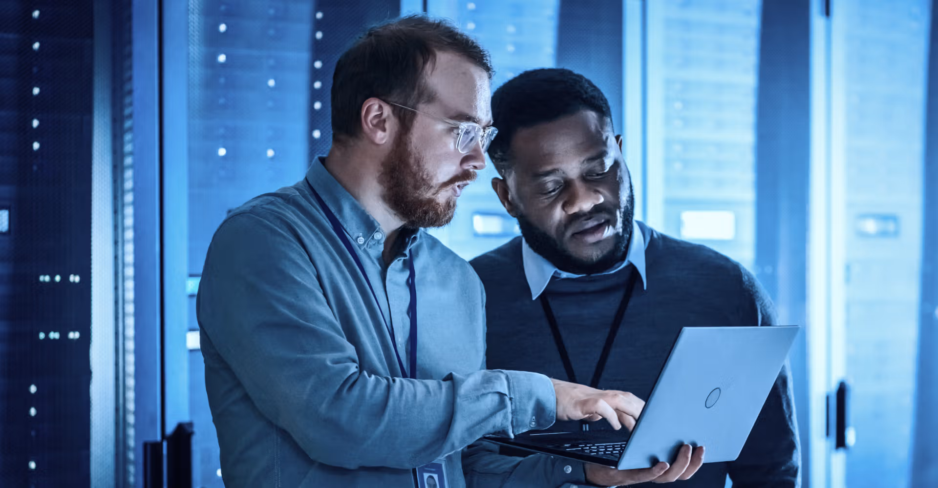 Two IT professionals examining data on a laptop in a server room with blue lighting.