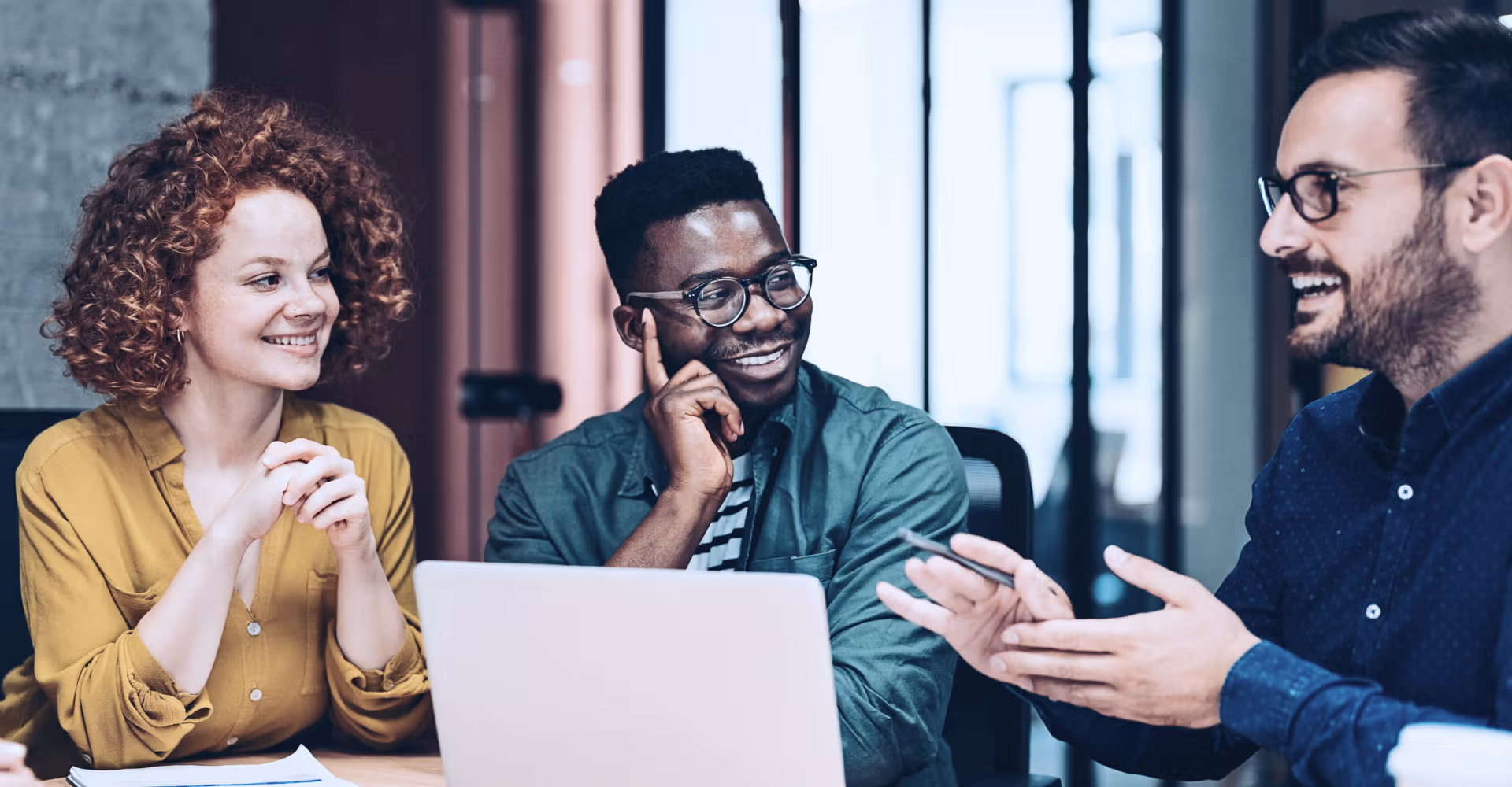 Three colleagues smiling and discussing work around a laptop in a modern office setting.