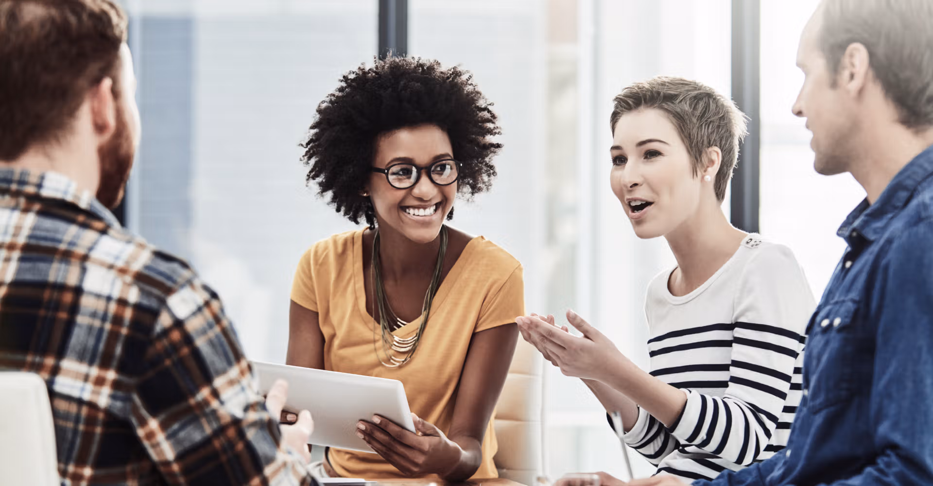 Four coworkers engaged in a lively discussion, one holding a tablet, in a bright office setting.