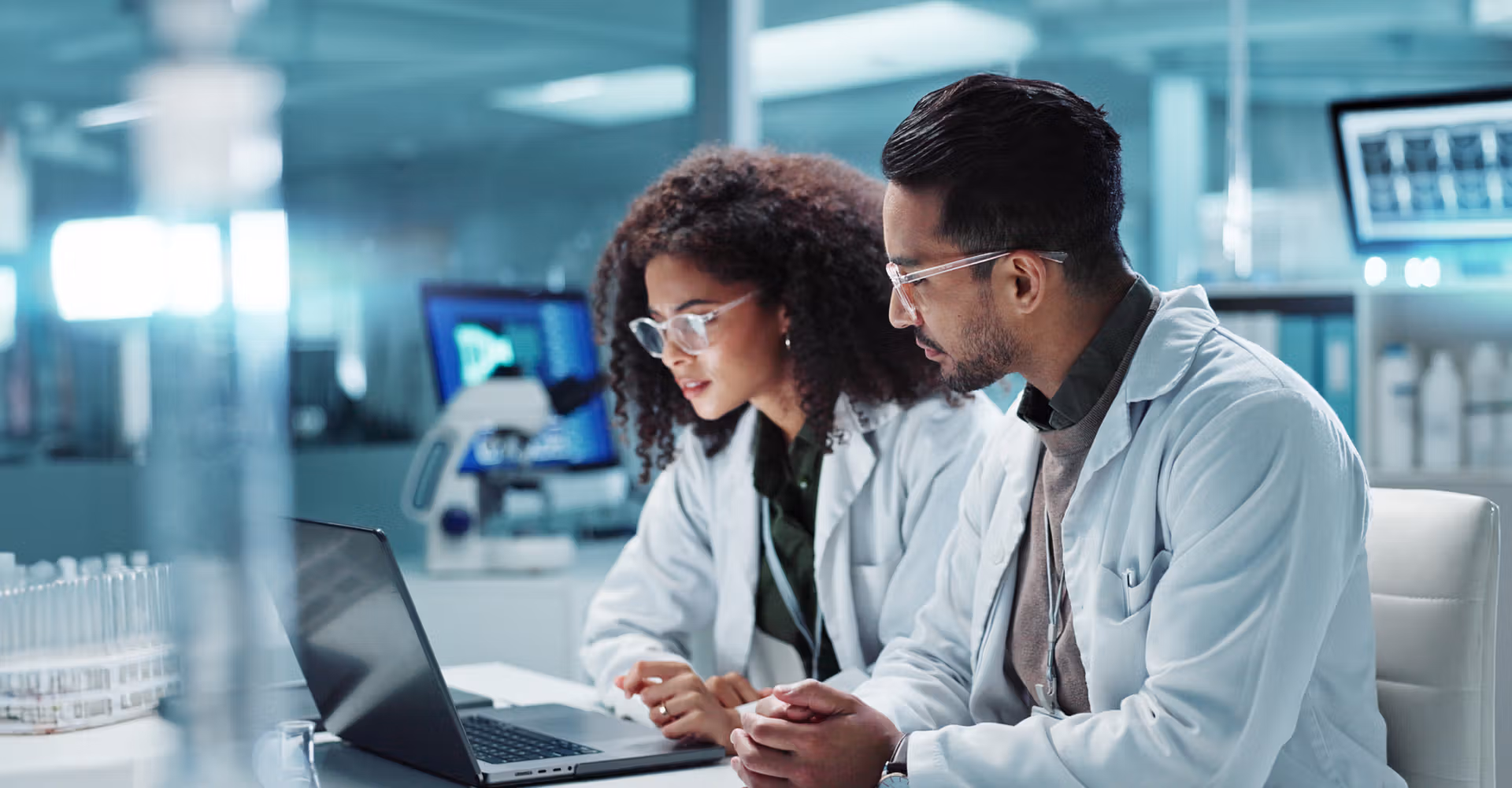 Two scientists in lab coats and glasses analyzing data on a laptop in a modern lab.