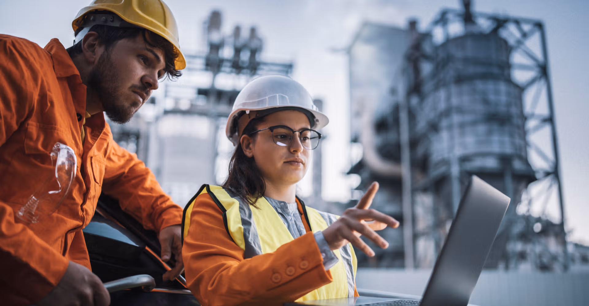 Two industrial workers in helmets and safety gear using a laptop at a factory site.