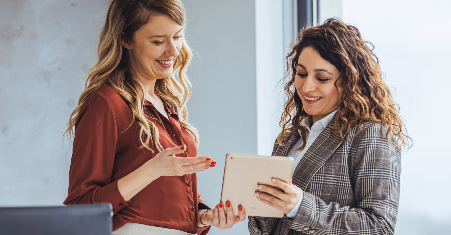 Two women smiling and discussing something while looking at a tablet in an office setting.