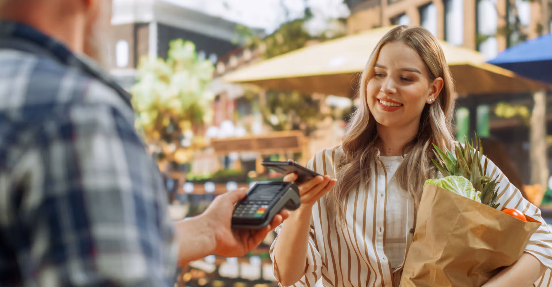 Smiling woman paying with phone using contactless terminal while holding grocery bag outdoors.