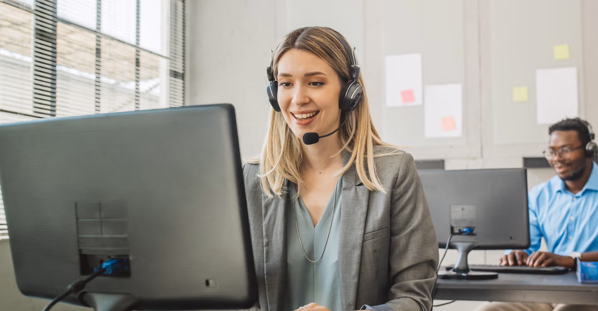 Smiling woman with headset using computer in office, man with headset working in background.