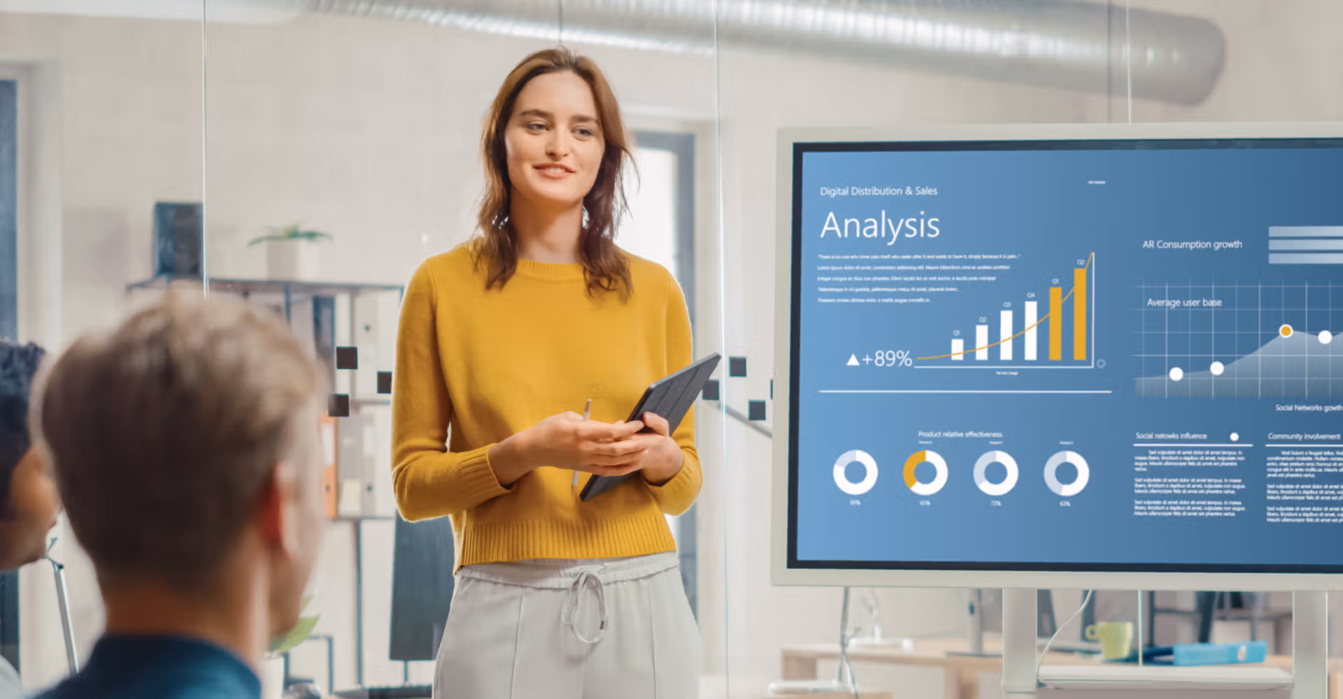 Woman in yellow sweater presenting digital sales analysis on screen to seated colleagues in office.