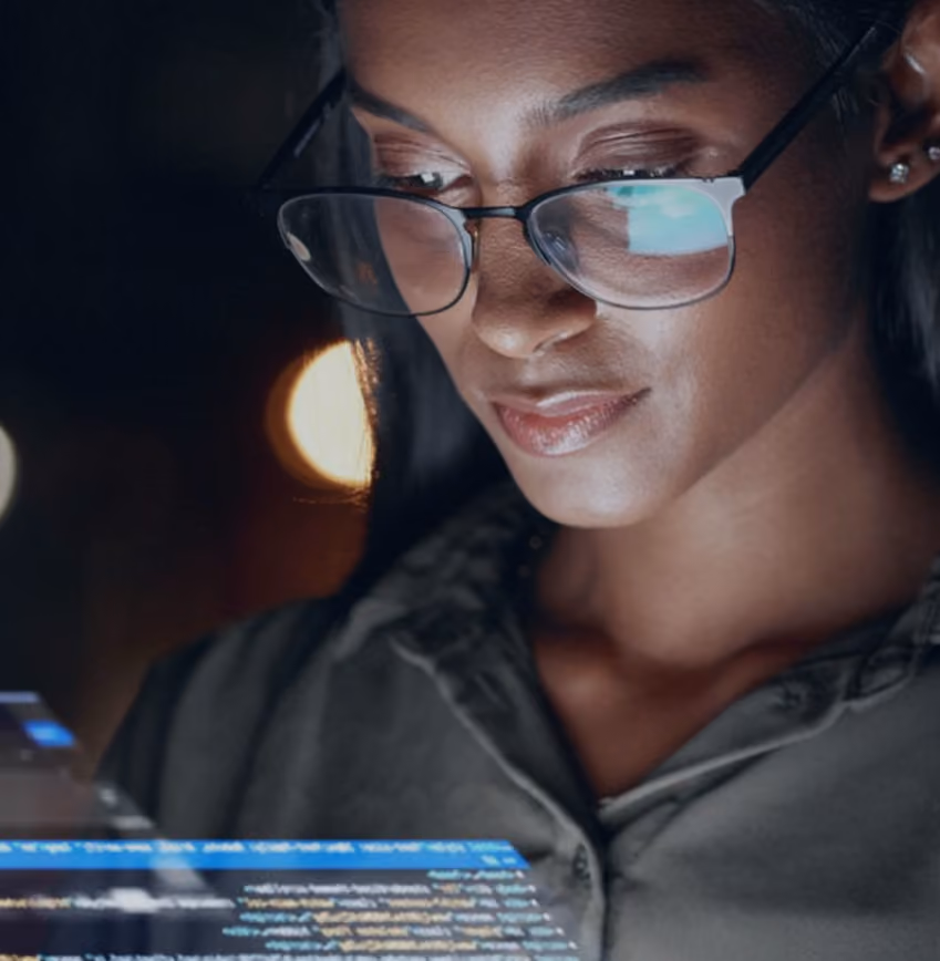 Woman wearing glasses focused on a holographic screen with code in a dark environment.