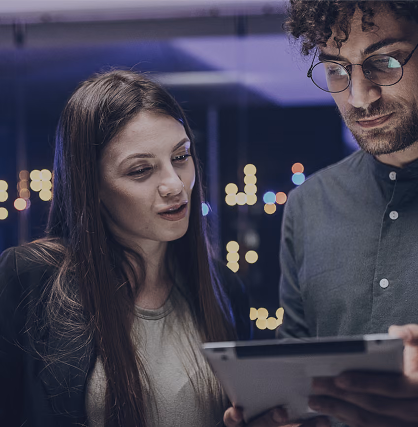 Two colleagues reviewing content on a tablet with blurred city lights in the background at night.