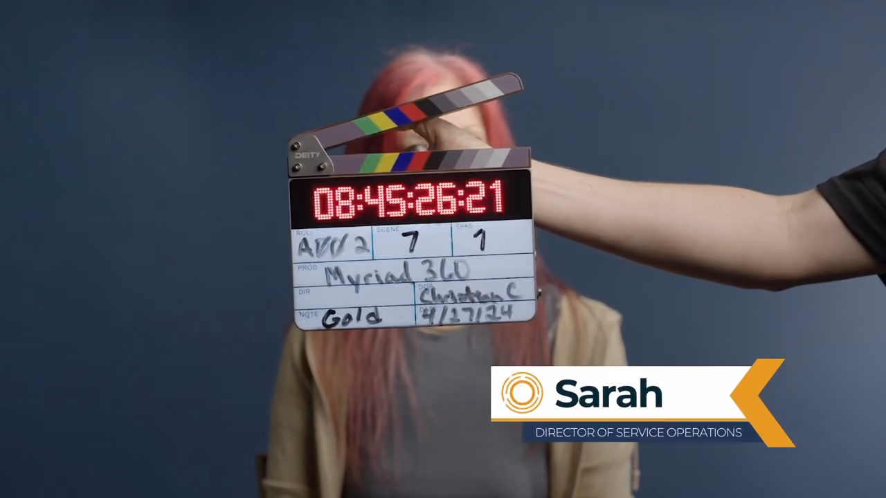 Clapperboard held in front of a woman with pink hair labeled Sarah, director of service operations.